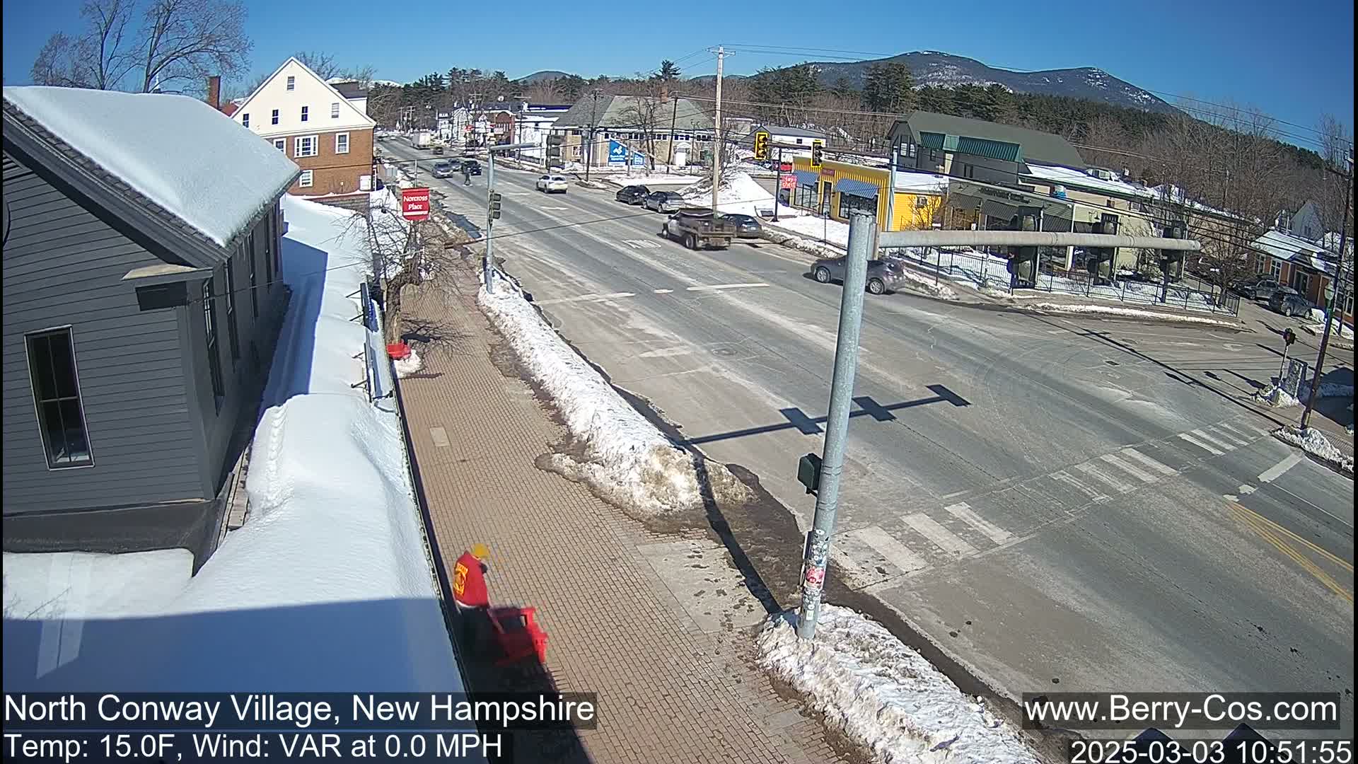 A sunny day shows a snow-covered street intersection in a village with a few cars driving and buildings on either side.