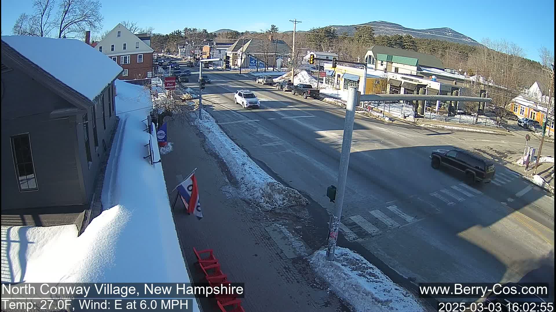 A sunny, snow-covered street scene in a village, with cars driving on a road that intersects another road.