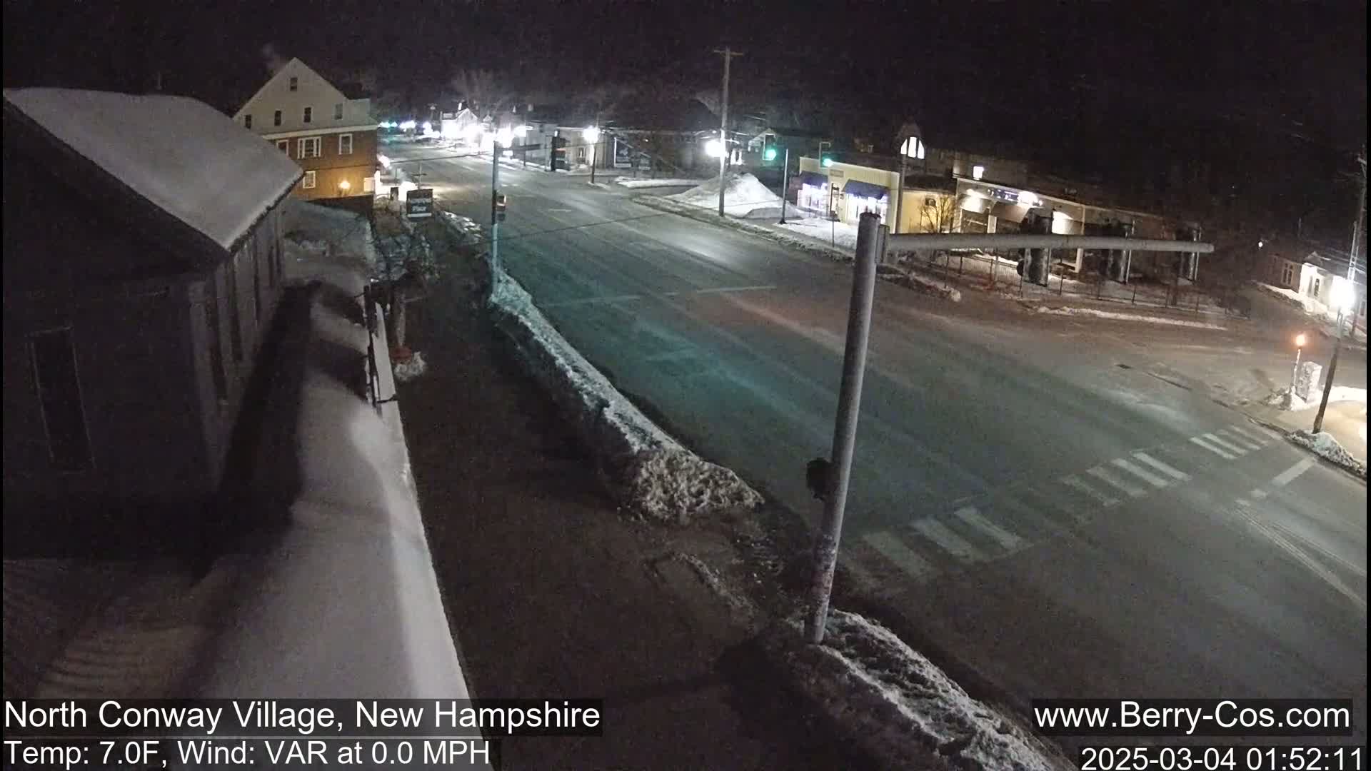 A snow-covered street intersection at night, with buildings and streetlights visible in a sparsely trafficked area.