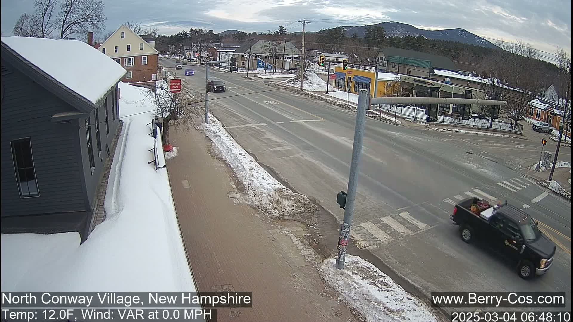 A snow-covered street scene in a village, with cars driving on a road that intersects with another, featuring buildings on either side and mountains in the background under a cloudy sky.