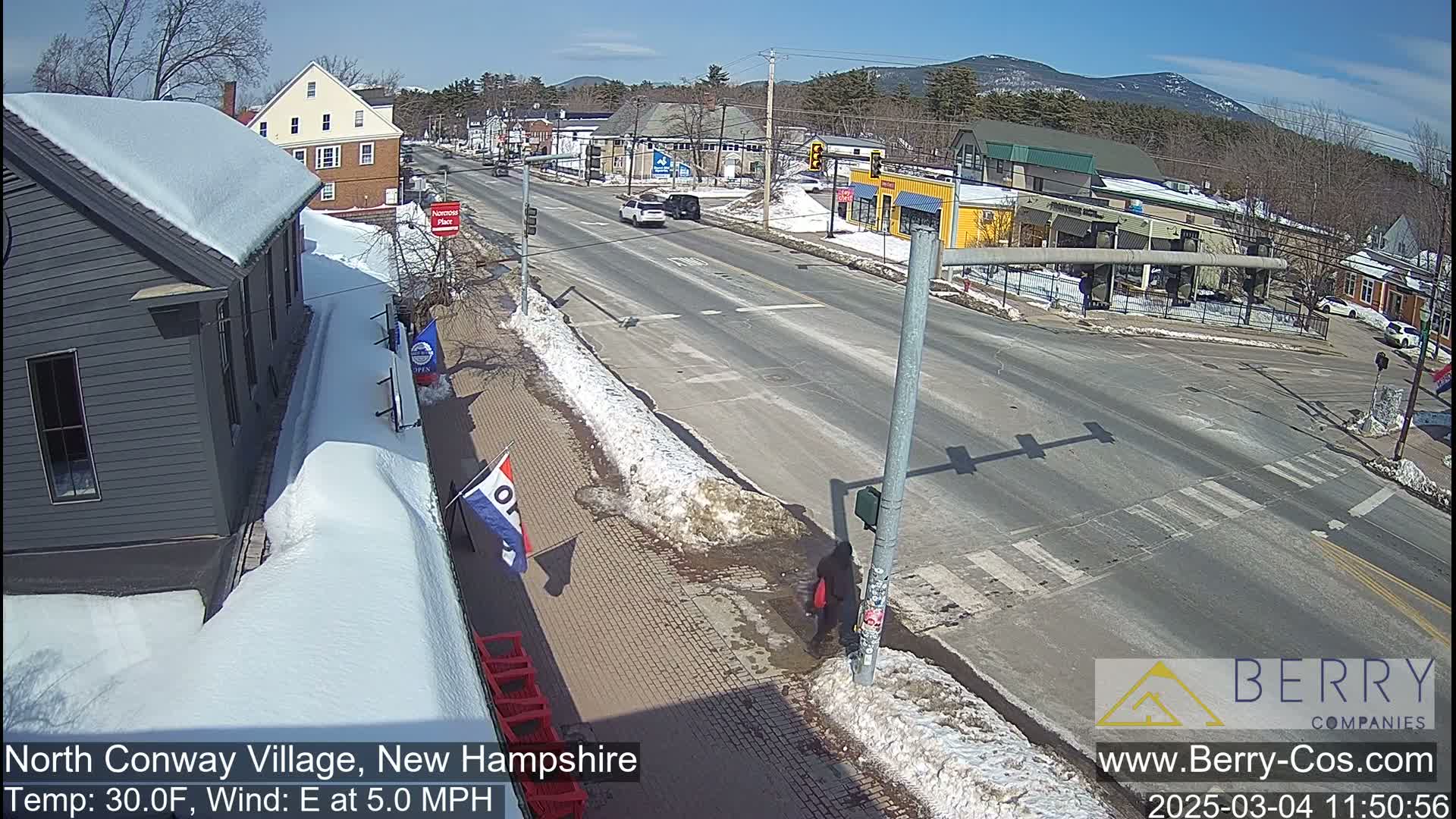 A sunny, snow-dusted street scene shows a mostly empty road with a few cars and a pedestrian, buildings with snow-covered roofs line the street, and mountains are visible in the background.