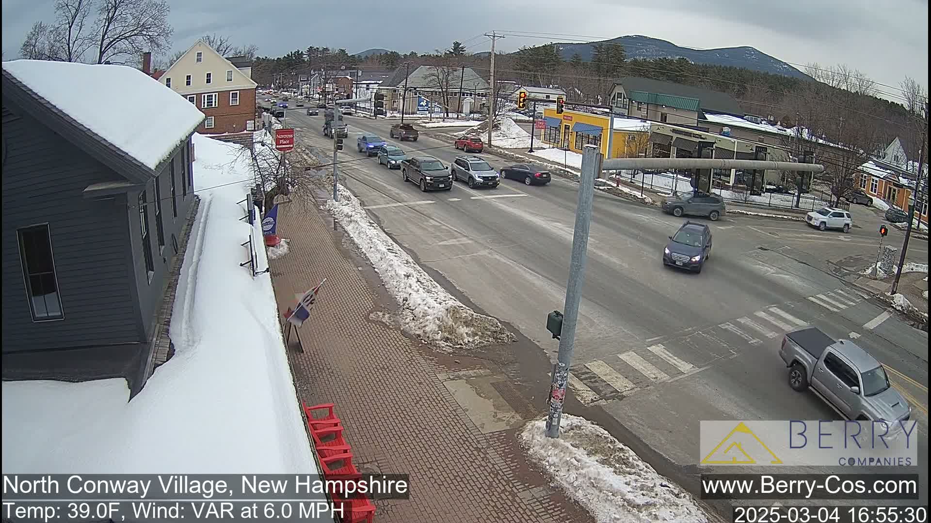 A snow-covered street scene shows several cars driving on a road that intersects with another road, with partially snow-covered sidewalks and buildings along the sides, under an overcast sky.