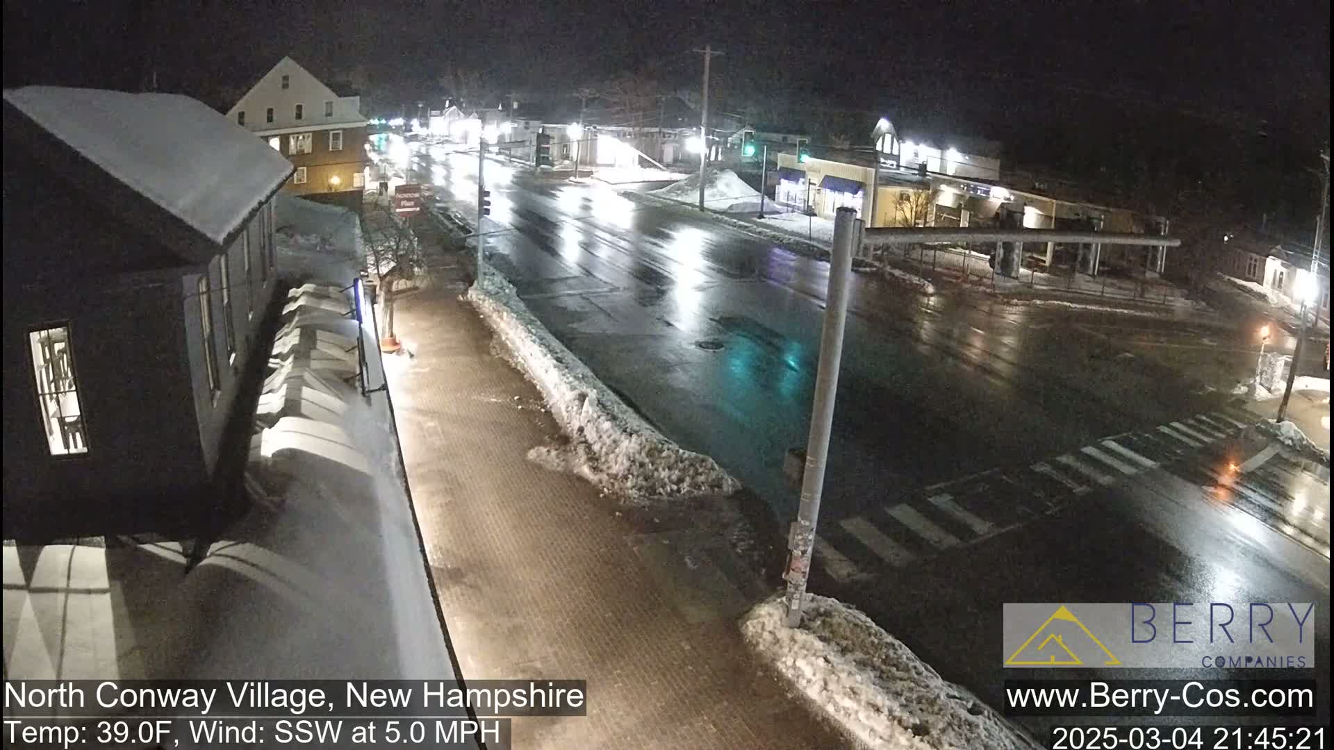 A wet, nighttime street scene shows a sparsely trafficked intersection with snow banks lining the sides of the road and buildings visible in the background under a clear sky.