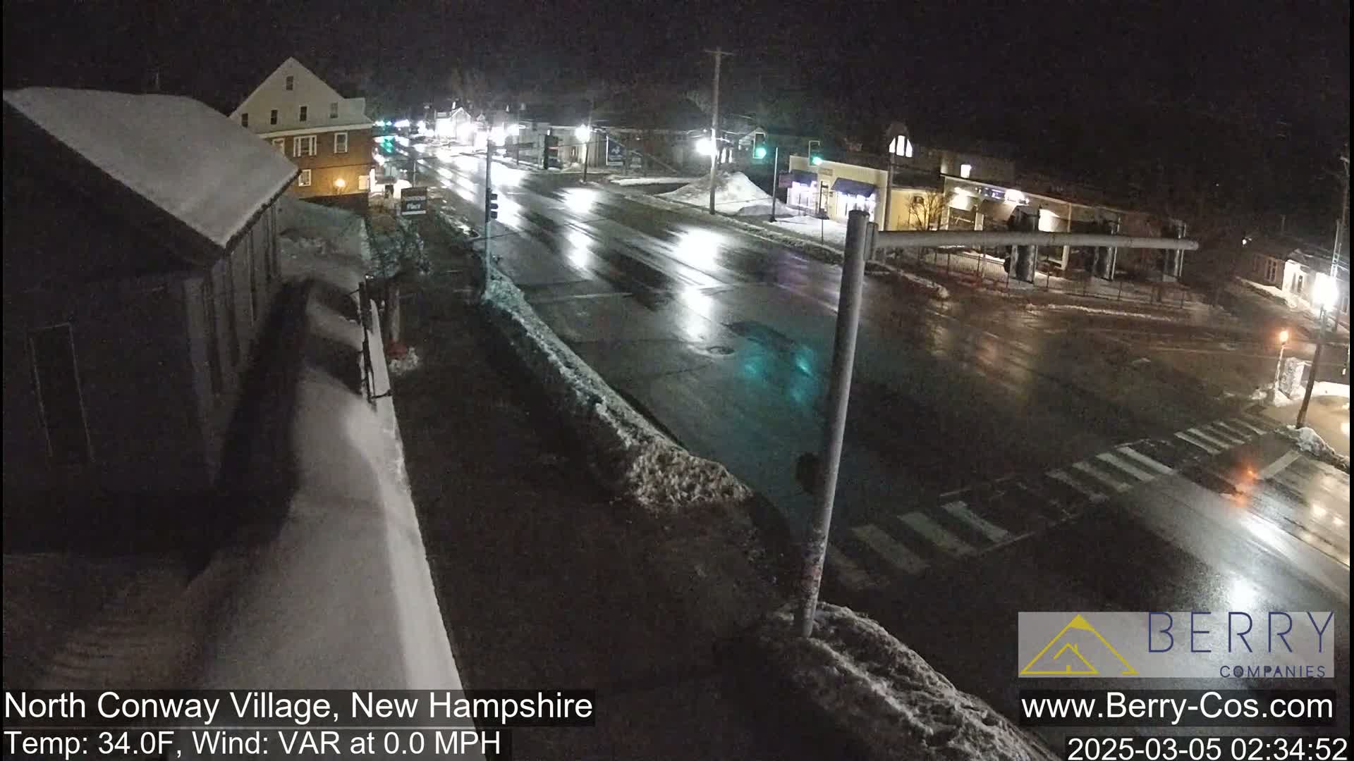 A nighttime view of a snow-covered street intersection with wet roads and sparsely-lit buildings under a dark sky.