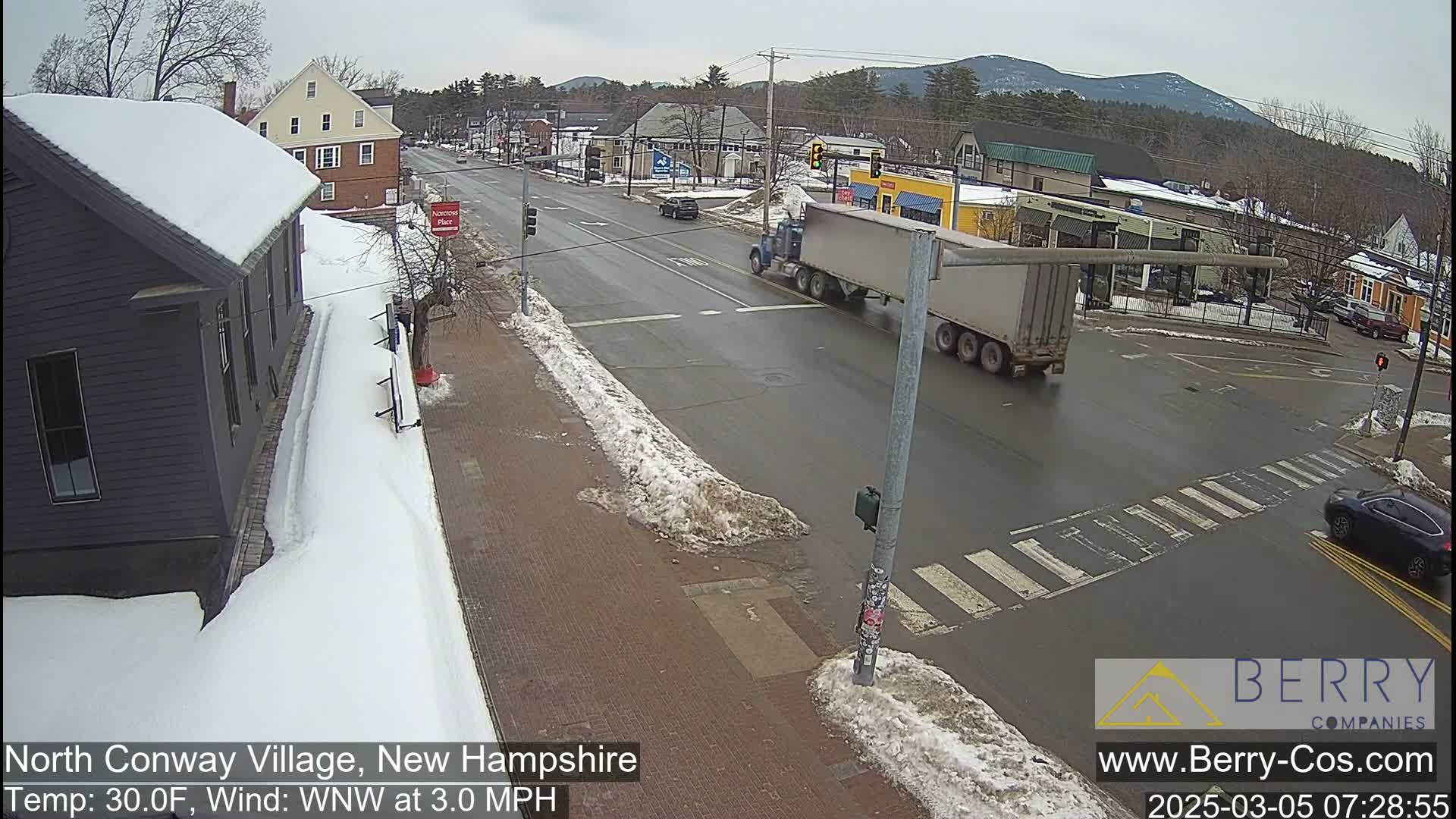 A snow-covered street scene in a small town shows a truck and several cars at an intersection under an overcast sky.