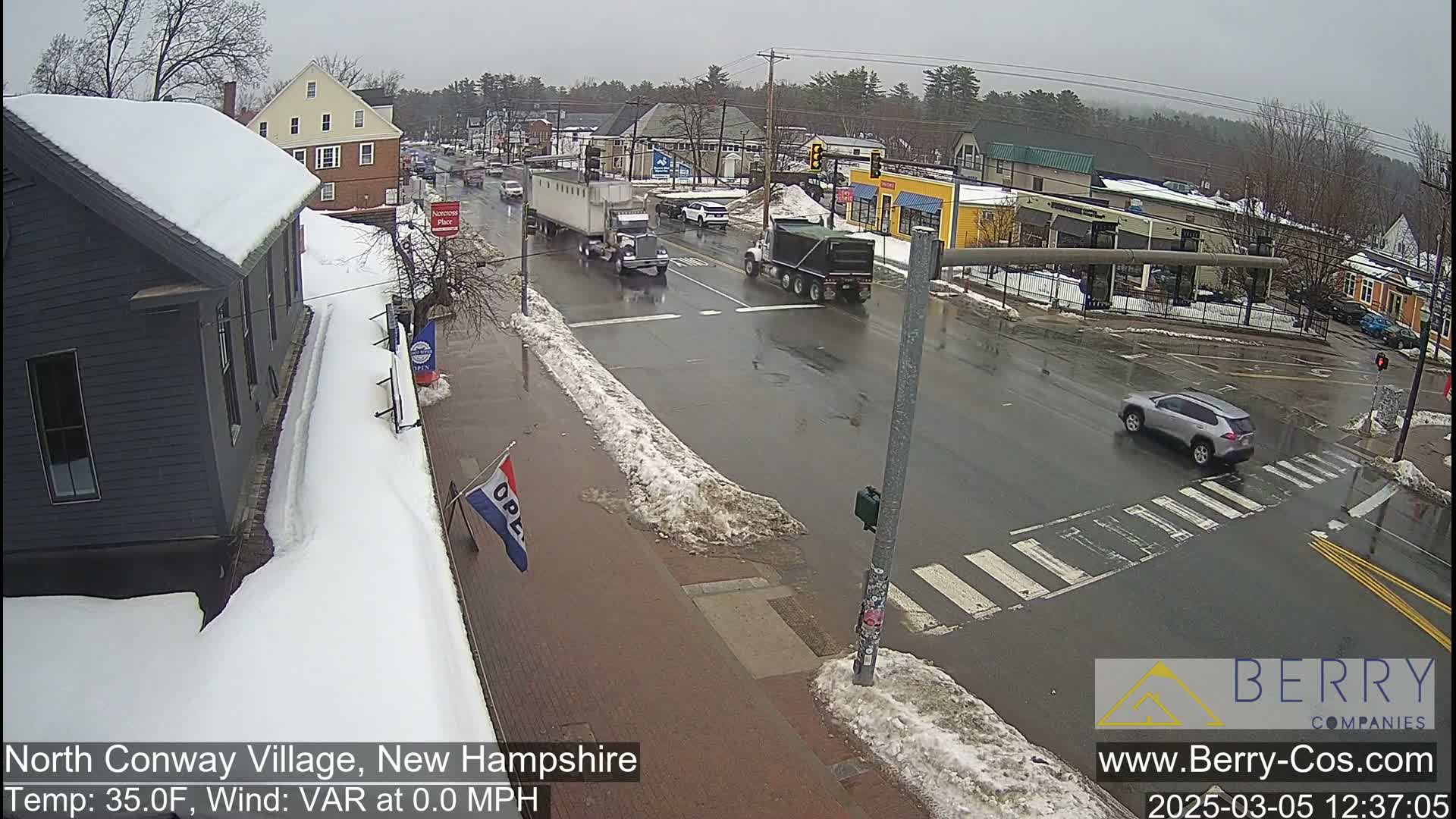 A wet road intersection with light traffic, snow on the sidewalks, and overcast skies.