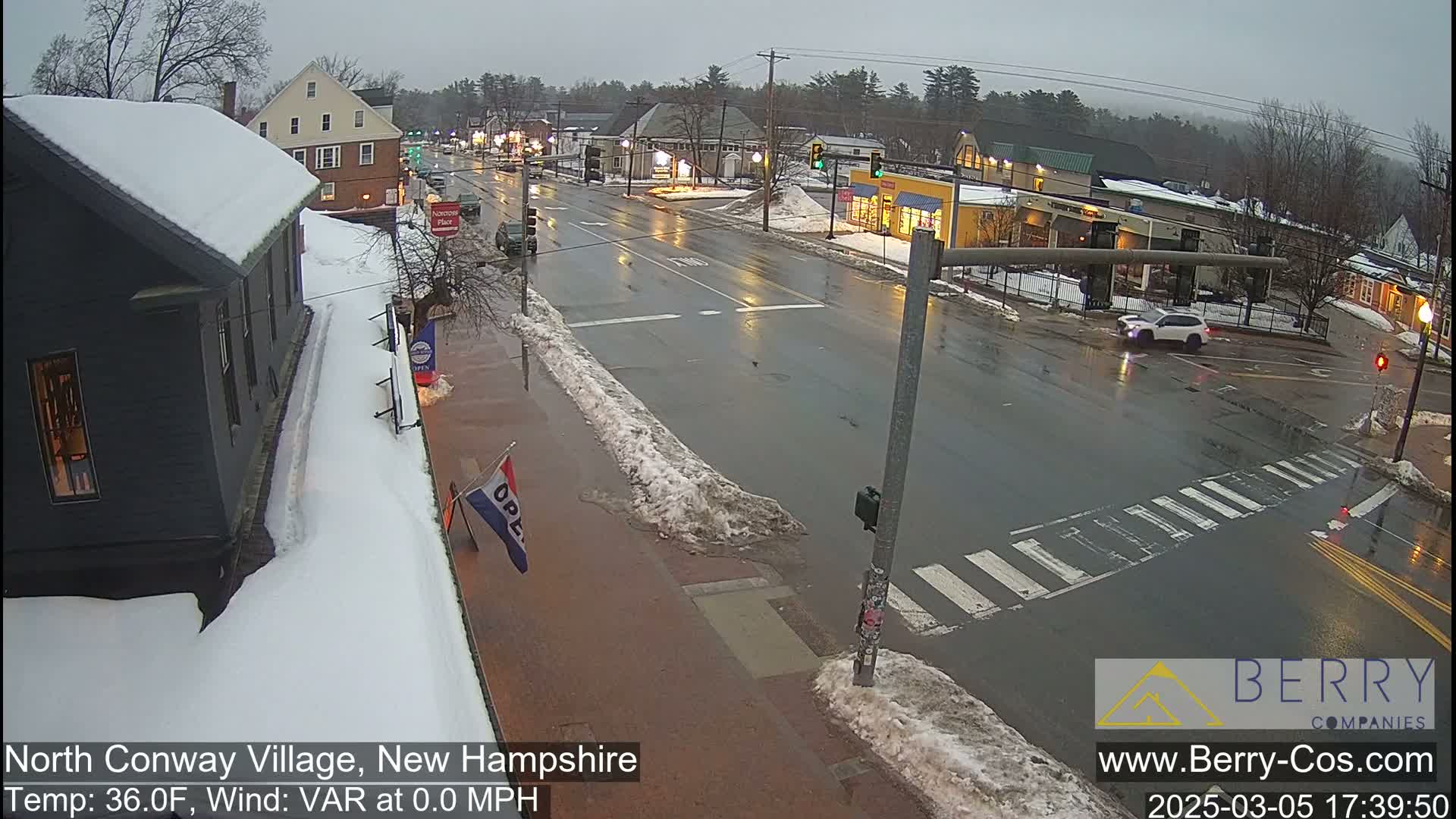 A wet, overcast street scene shows a village intersection with snow banks lining the sides of the road and some snow on building rooftops.
