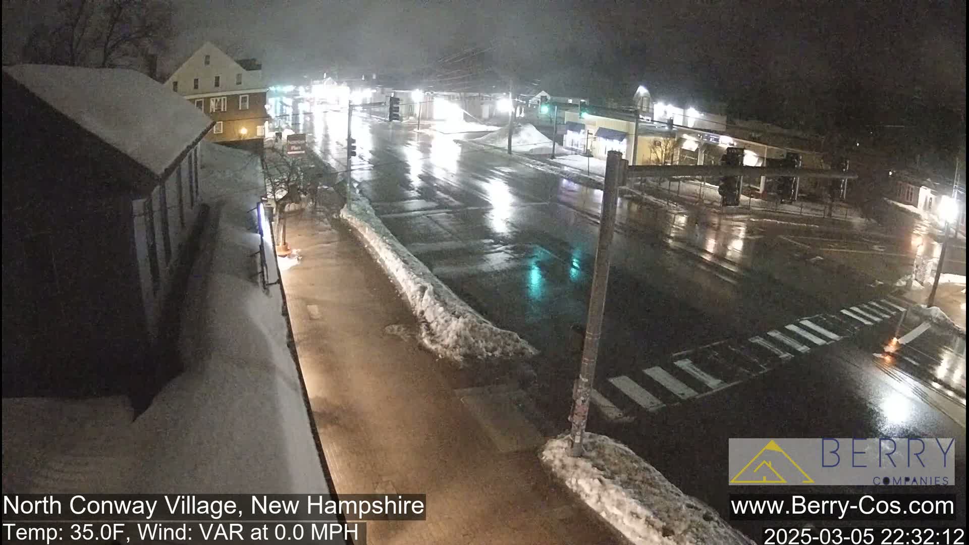A wet, nighttime street scene shows a few cars on a road bordered by snow banks and buildings under dim streetlights.