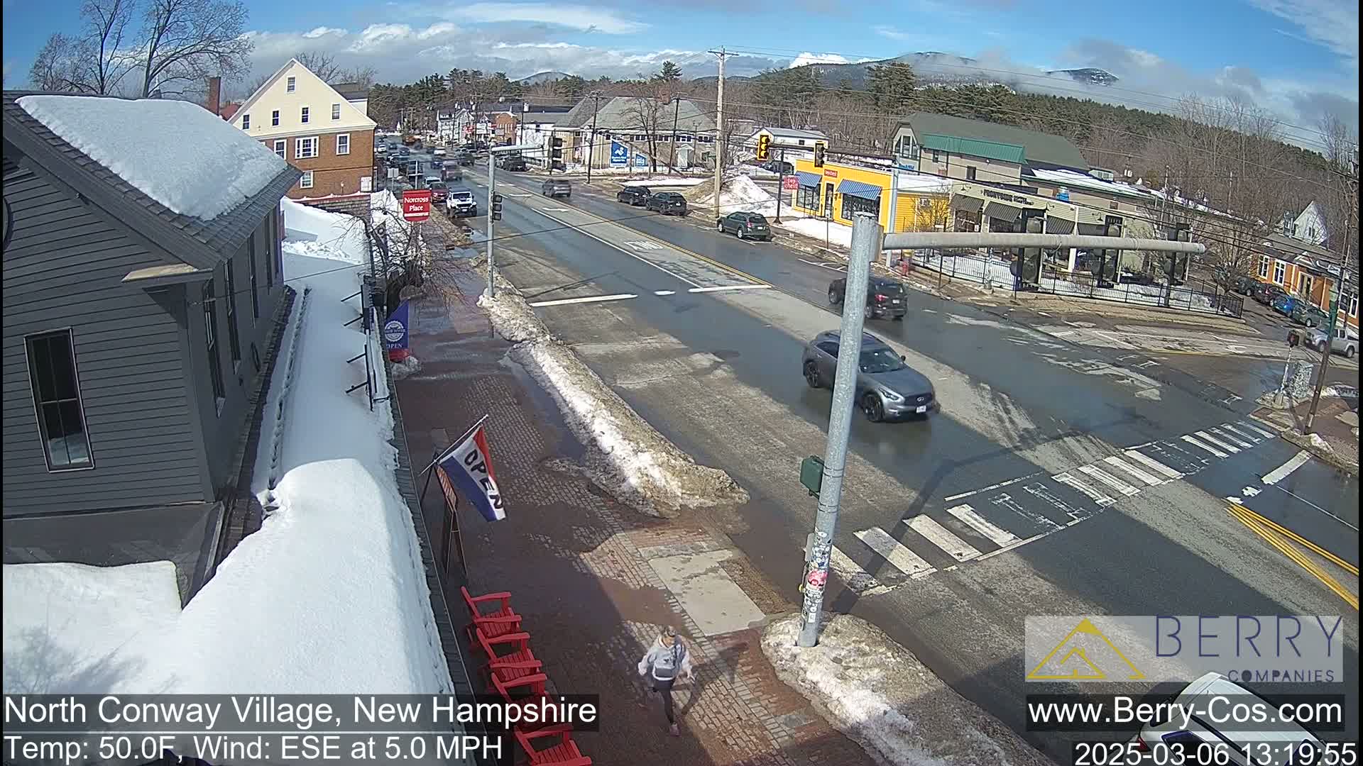 A sunny day in a snow-covered village with cars driving on a road and a pedestrian walking on the sidewalk.