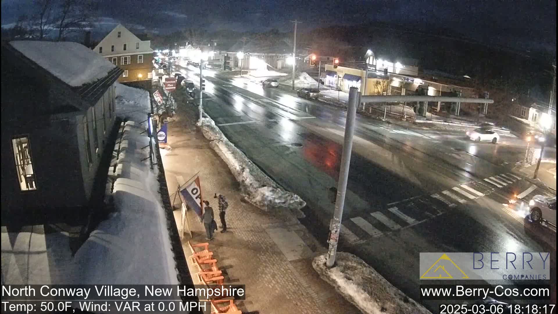 A wet, nighttime street scene shows several cars driving past buildings with snow on the ground and rooftops, under a dark sky.