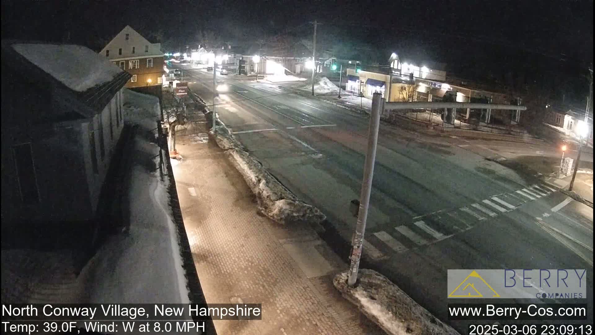 A snow-covered nighttime street scene shows a multi-lane road intersection with several buildings and some cars.