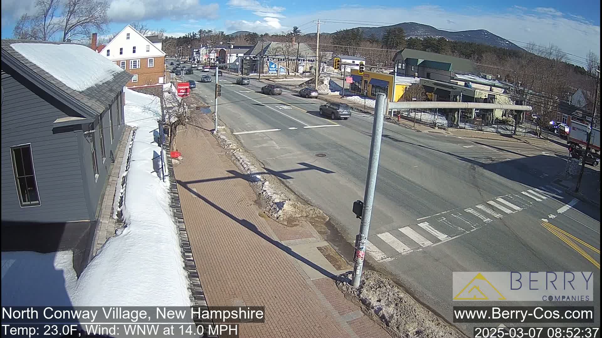 A sunny day with snow on the ground shows a street intersection in a small town with several cars, buildings, and some patches of bare ground.