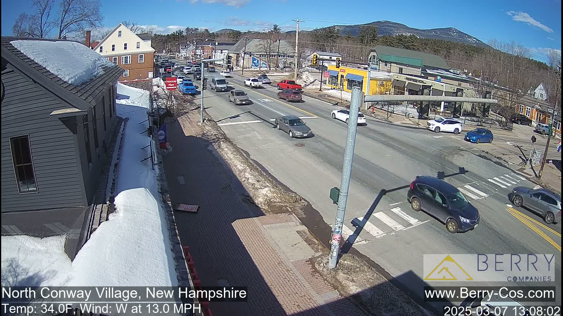 A sunny, wintery street scene shows several cars at an intersection, with snow visible on the sidewalks and some remaining on building roofs.