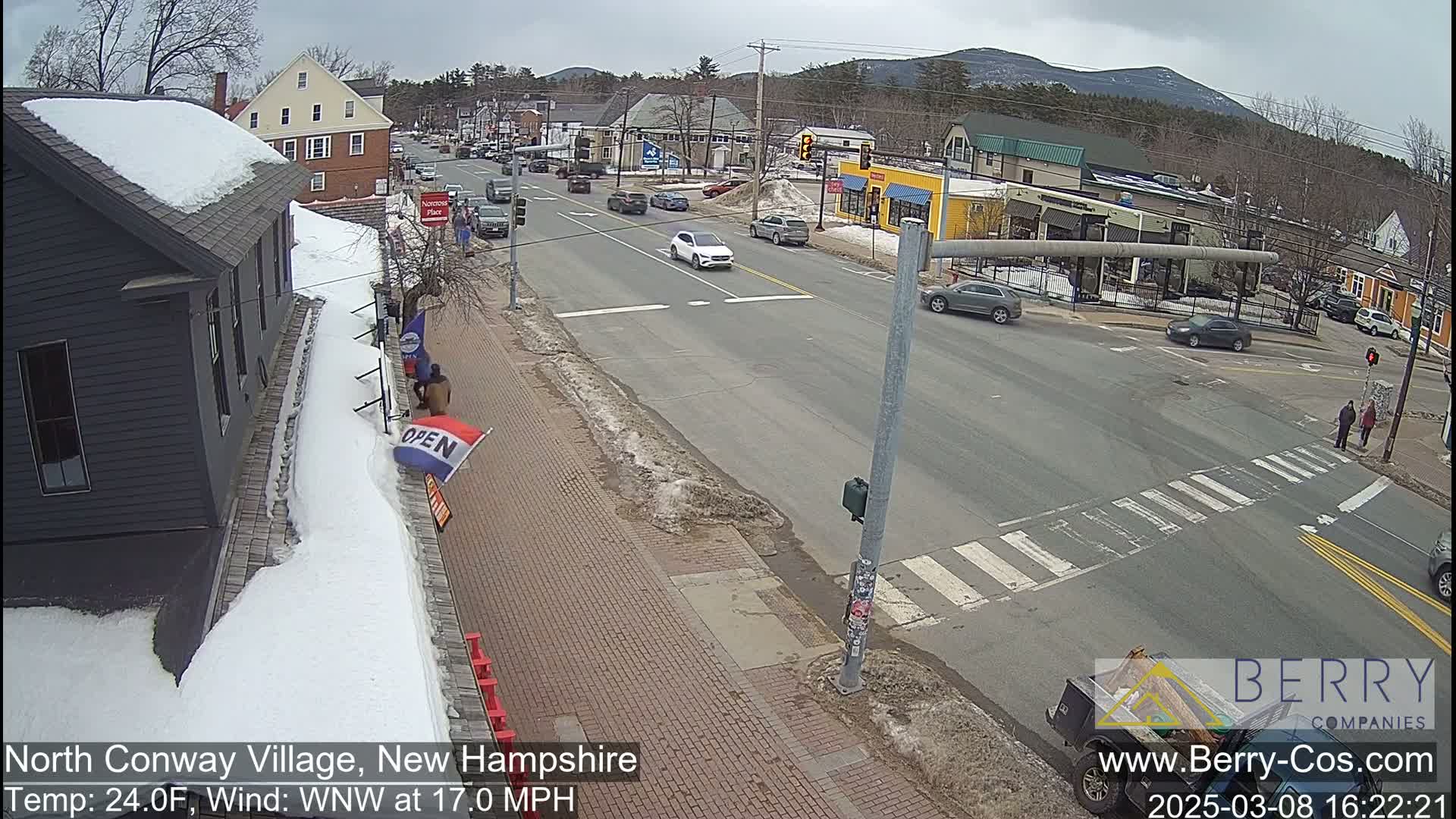 A snow-covered street scene in a small town shows cars at an intersection on a partly cloudy day.
