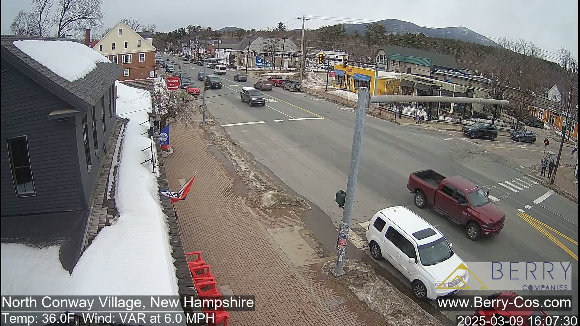A street scene shows several vehicles driving on a road lined with snow-covered sidewalks and buildings, under a partly cloudy sky.