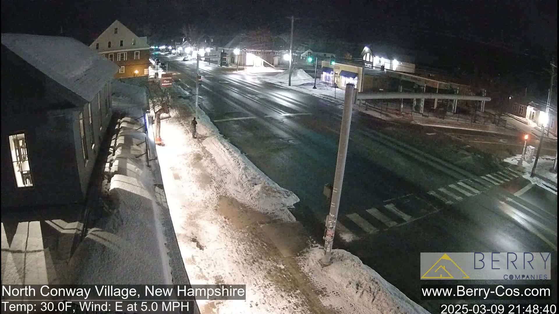A snow-covered street intersection at night, with a few buildings and a pedestrian visible under streetlights.