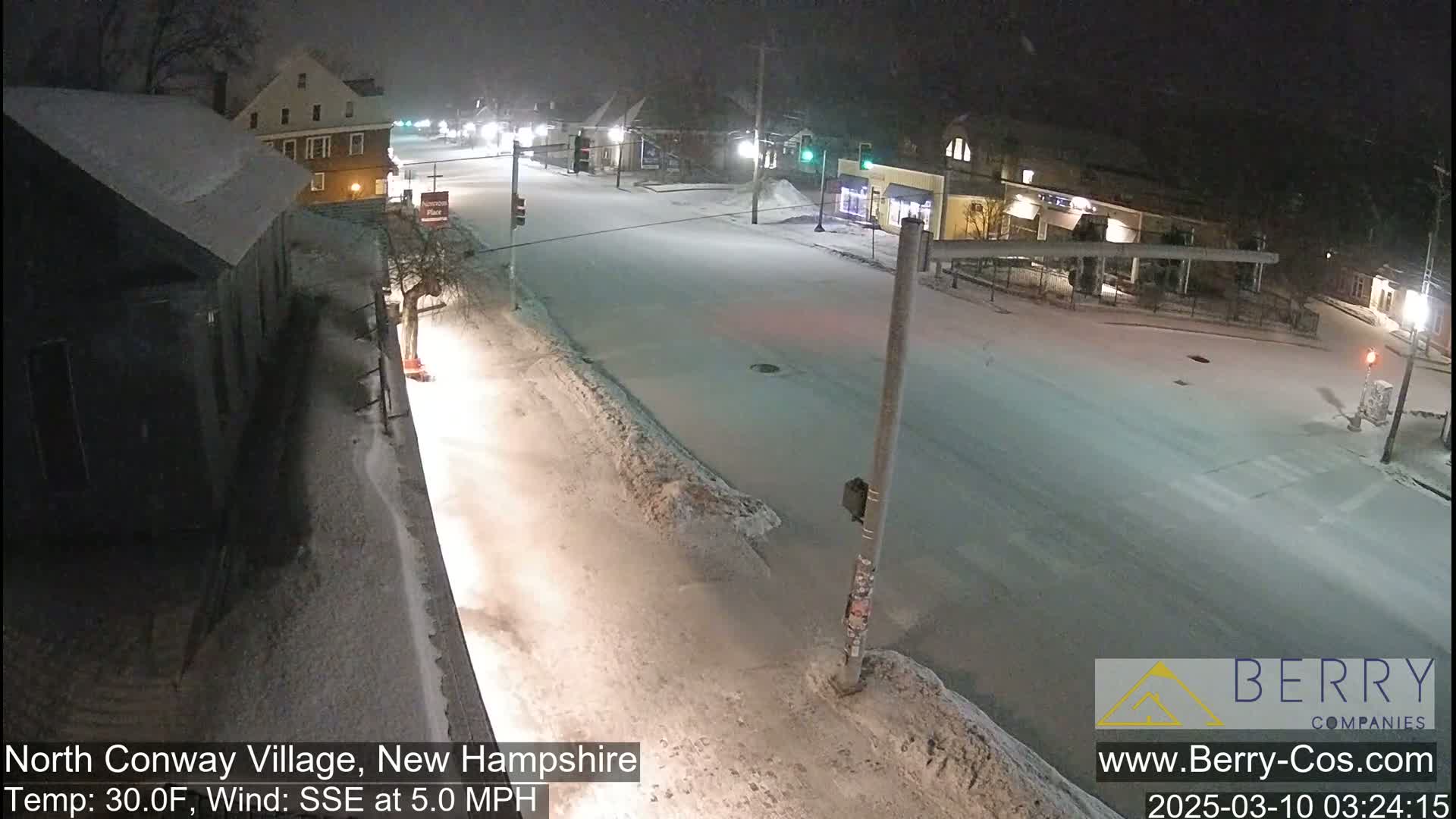 A snow-covered street scene at night, illuminated by streetlights, with buildings and some light traffic visible under a lightly snowing sky.