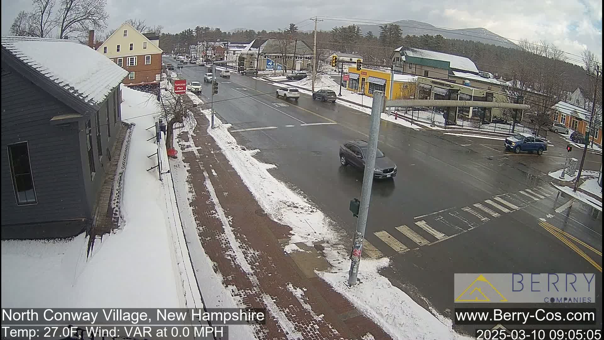 A snow-covered street scene shows several vehicles at an intersection, with buildings and some bare trees in the background under a cloudy sky.