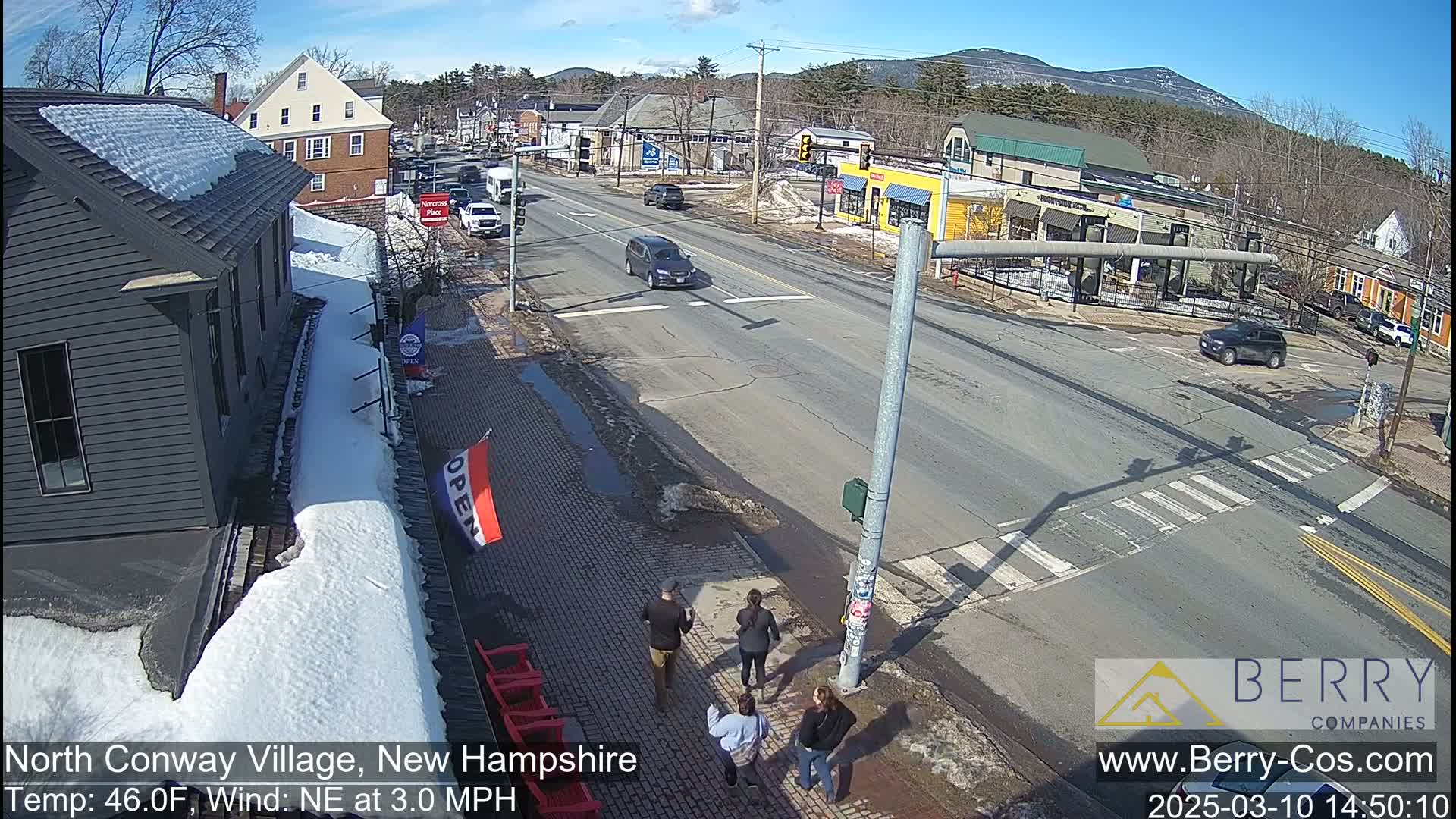 A sunny day in a snow-dusted village shows a street intersection with several cars and pedestrians, and mountains in the background.