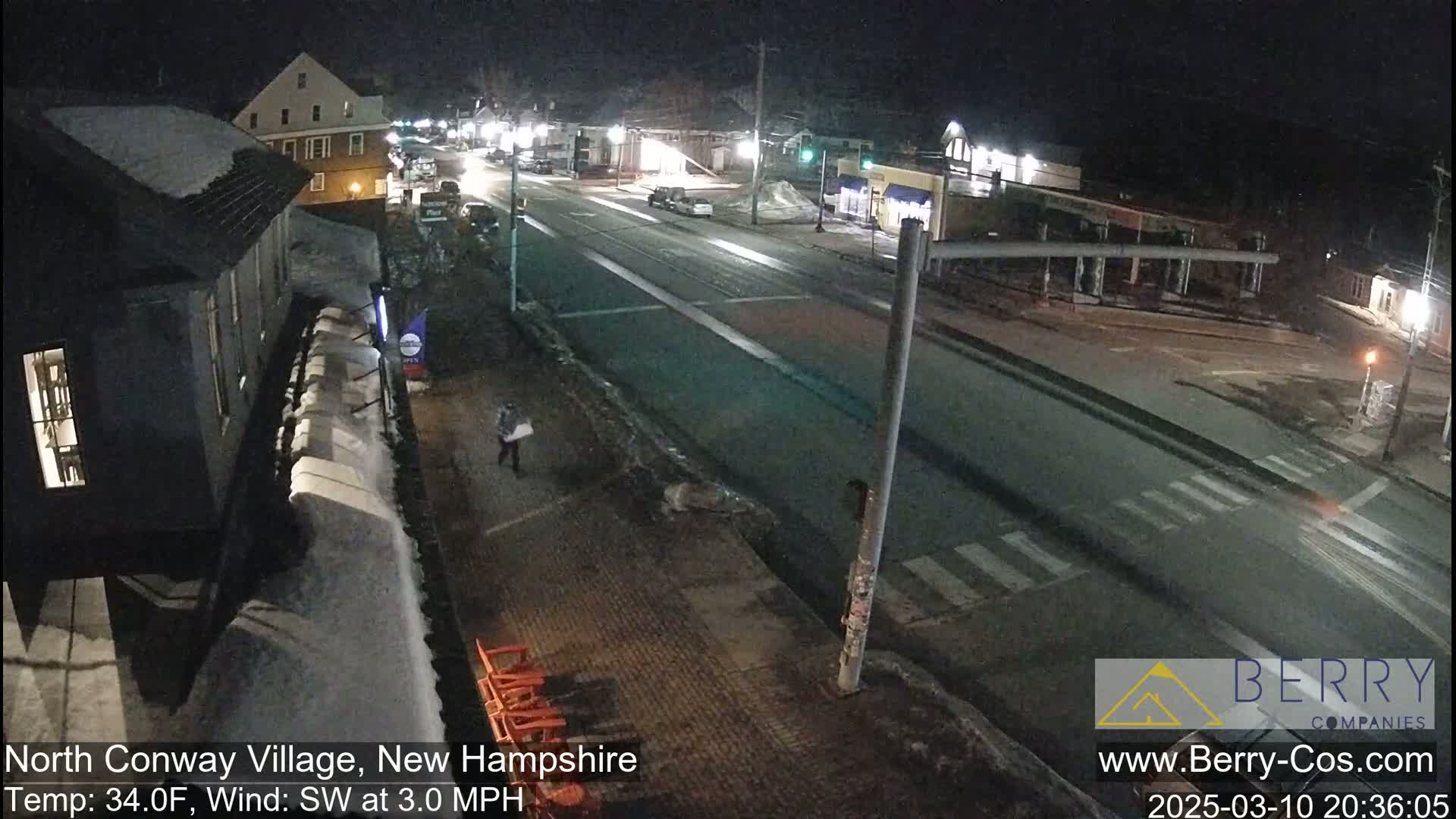 A nighttime street scene shows a sparsely traveled road with some cars, buildings covered in snow, and a person walking along the sidewalk under a clear sky, with a southwest wind at 3.0 mph and a temperature of 34°F.