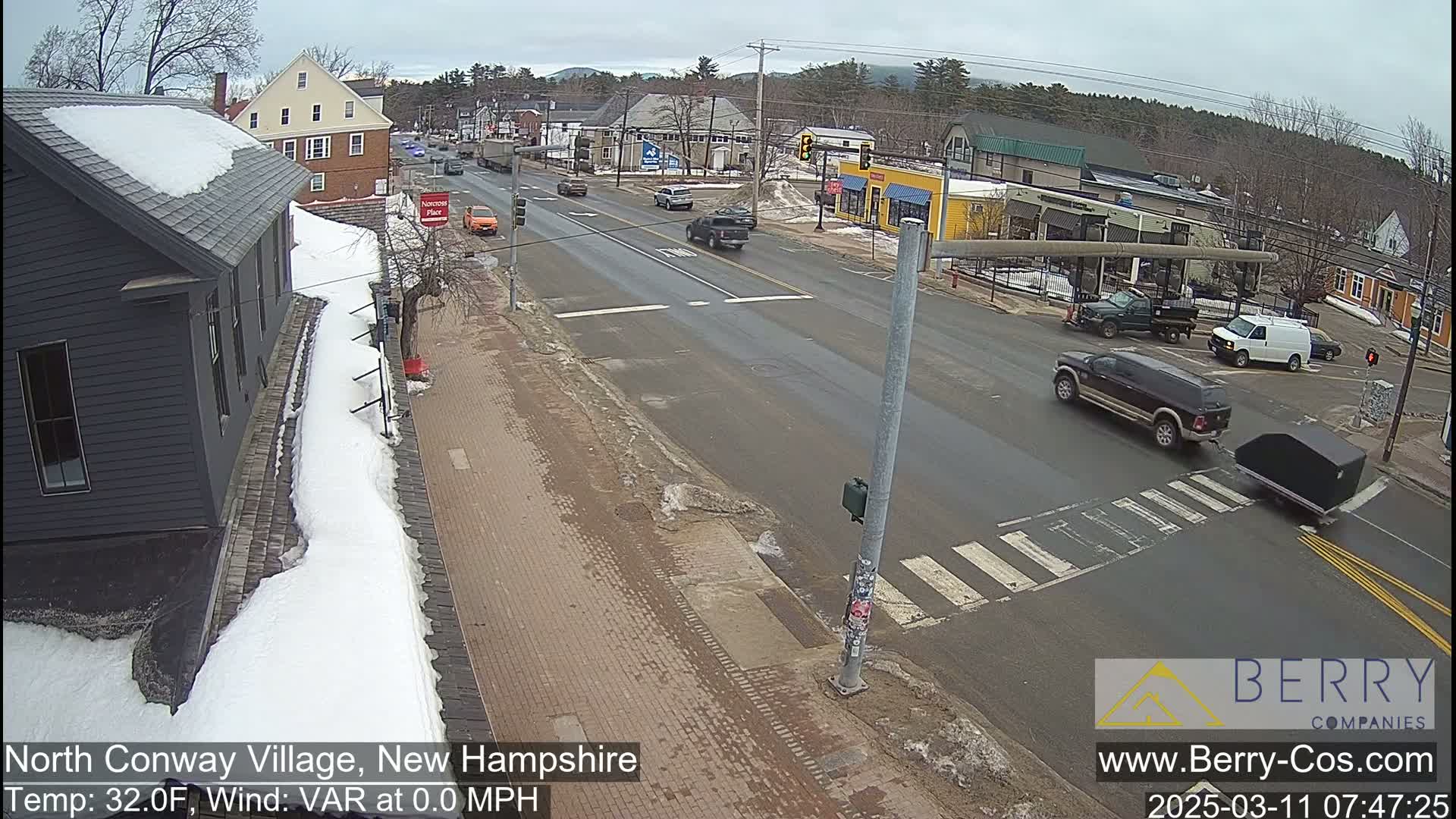 A snow-covered village street scene with several cars at an intersection under a cloudy sky.