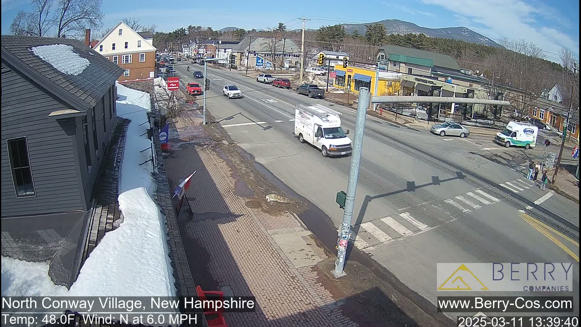 A sunny street scene shows several vehicles driving on a road that passes in front of various buildings, with some snow and ice still present on the sidewalks.