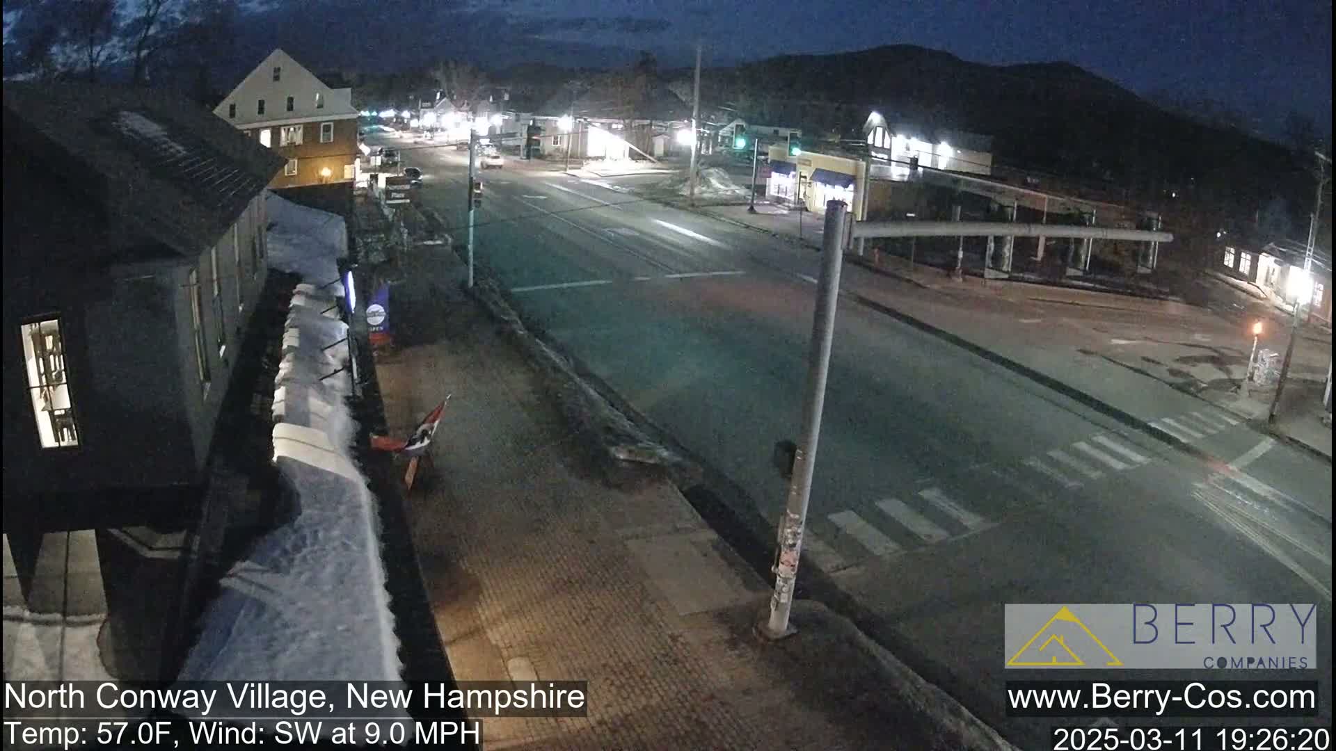 A nighttime view of a mostly empty road in a small town, with some snow on the ground and a southwest wind.