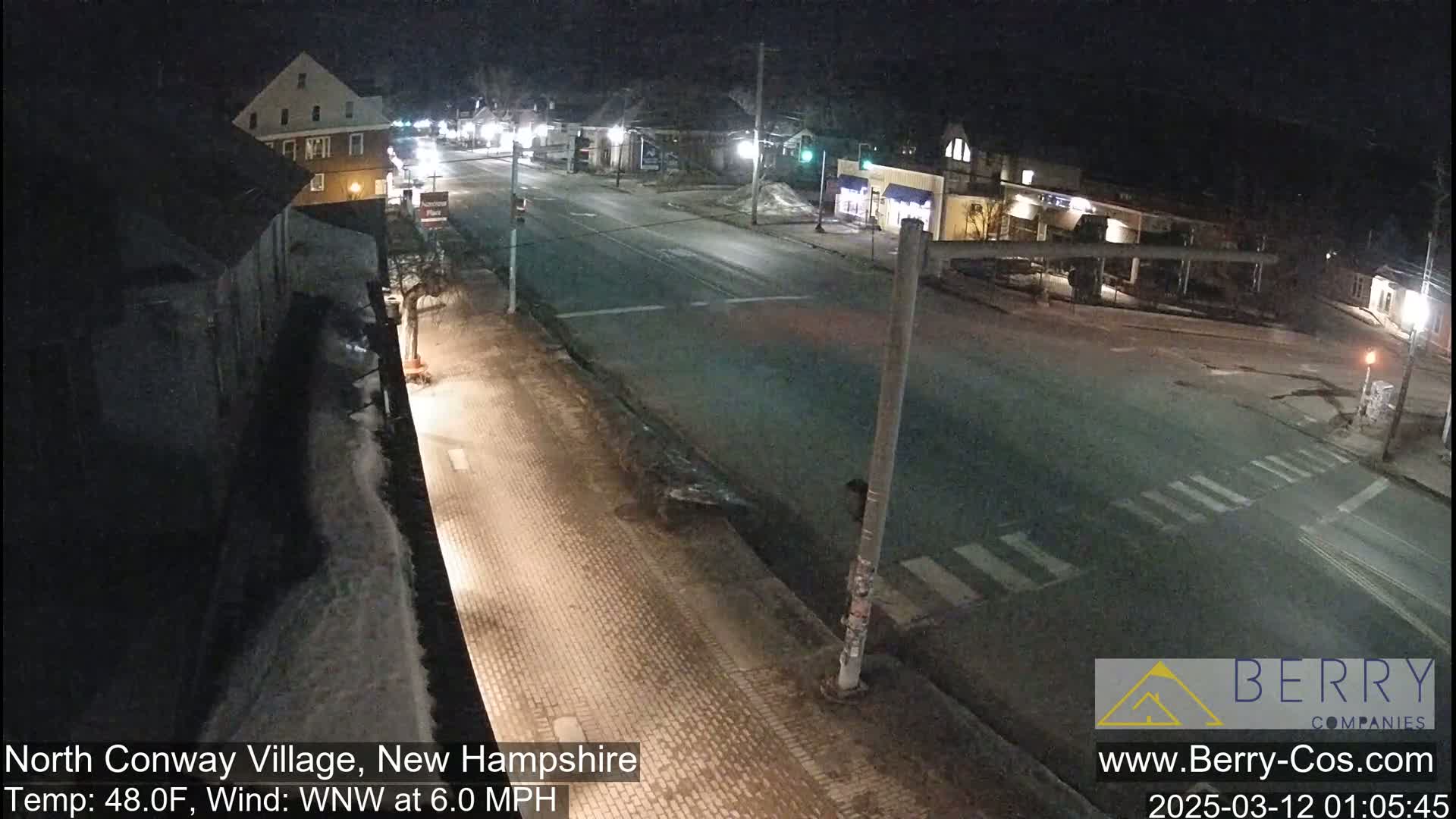A nighttime street scene shows a snow-covered street with buildings and minimal traffic under partly cloudy skies, with a temperature of 48°F and a west-northwest wind at 6.0 mph.