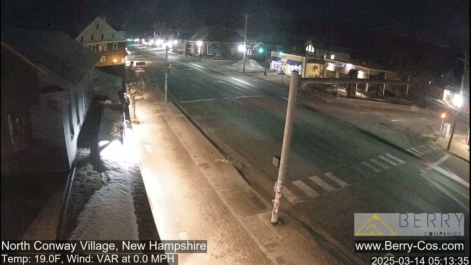 A snow-covered street scene at night, showing a quiet intersection with buildings on either side, under calm conditions.
