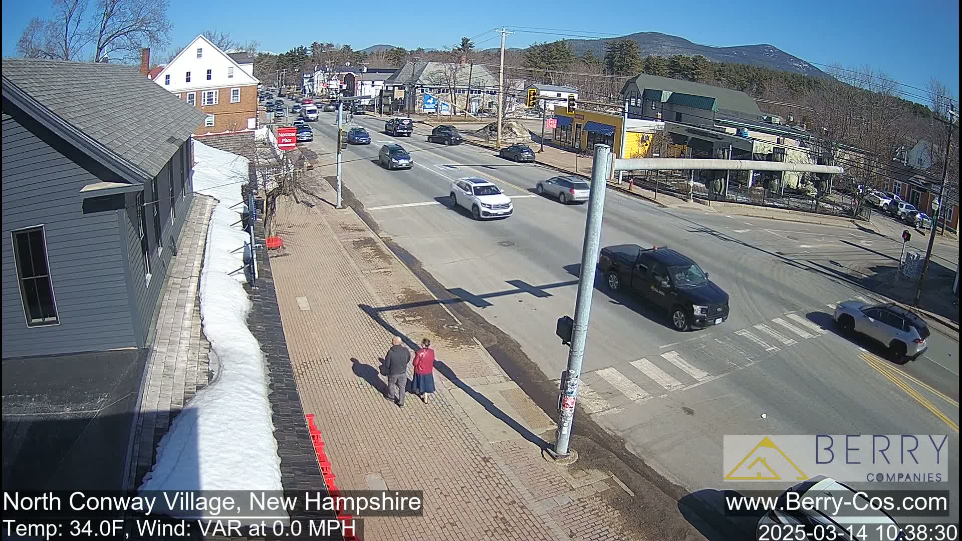 A sunny street scene shows a couple walking on a sidewalk beside a roadway with several cars, some snow on the ground, and distant mountains under a clear blue sky.