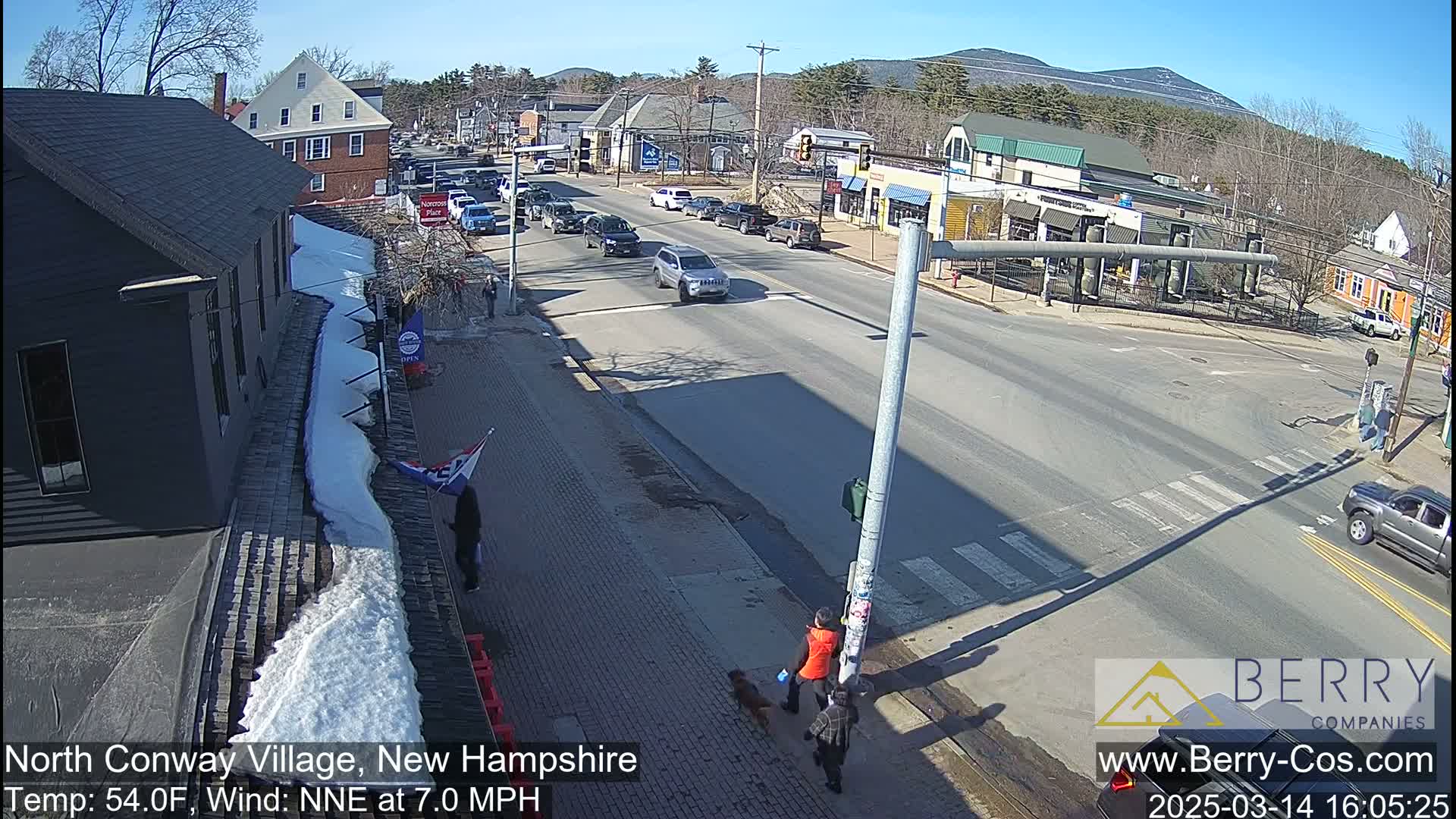 A sunny street scene shows a village intersection with cars and pedestrians, some snow on the ground near buildings, and mountains in the background.