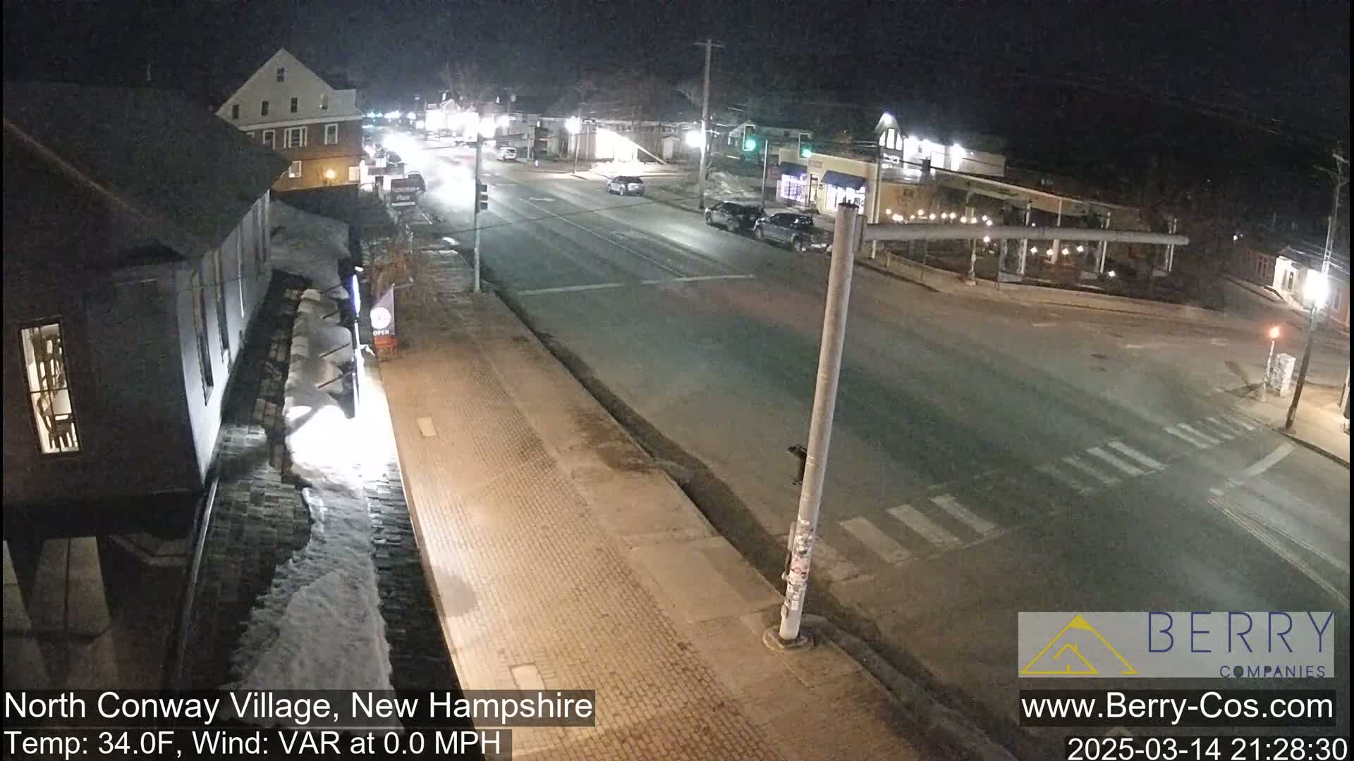 A nighttime street scene shows a mostly empty road intersection with a few cars, buildings lining the street, and patches of snow on the ground.