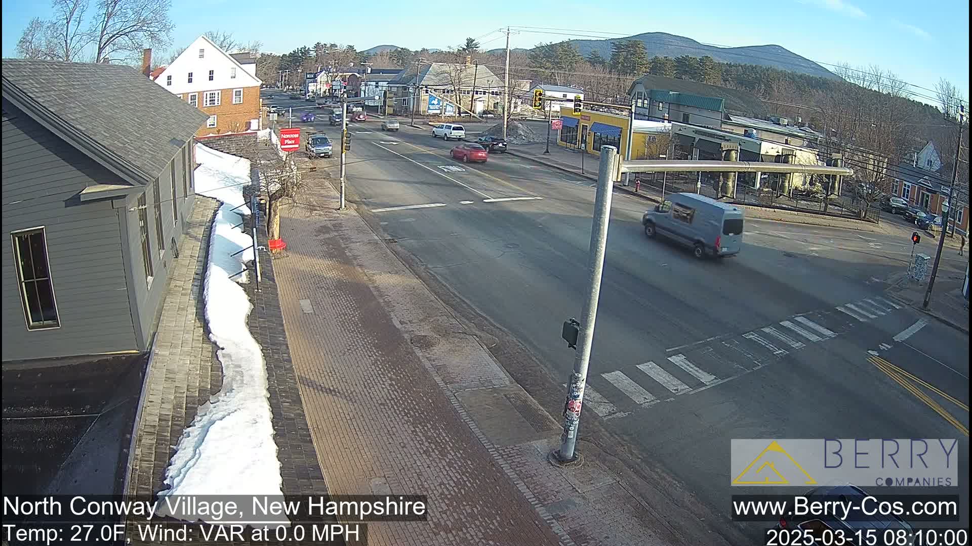A sunny street scene shows a sparsely trafficked intersection in a village setting, with some snow on the ground near buildings.