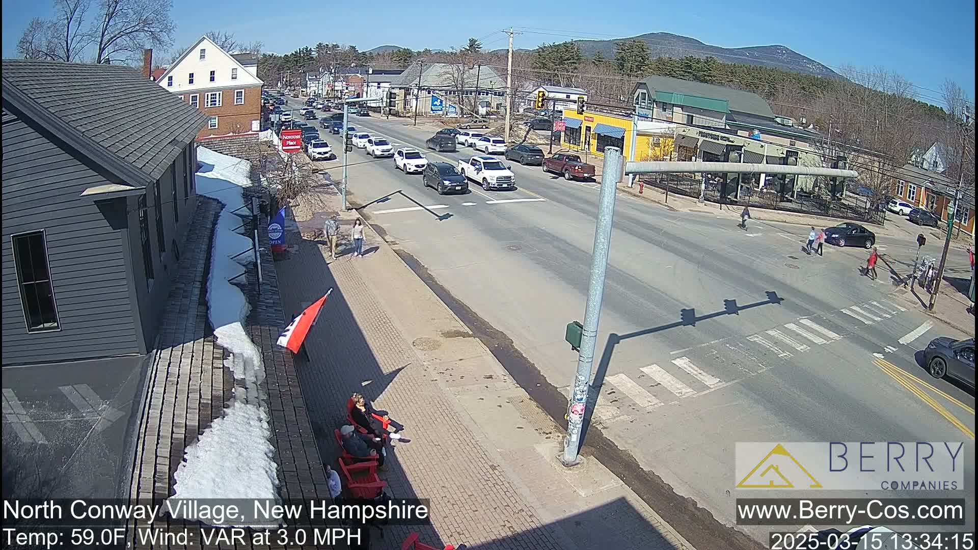 A sunny street scene shows several cars driving on a road, pedestrians crossing at a crosswalk, and some people sitting outside a building; melting snow is visible on the sidewalk.
