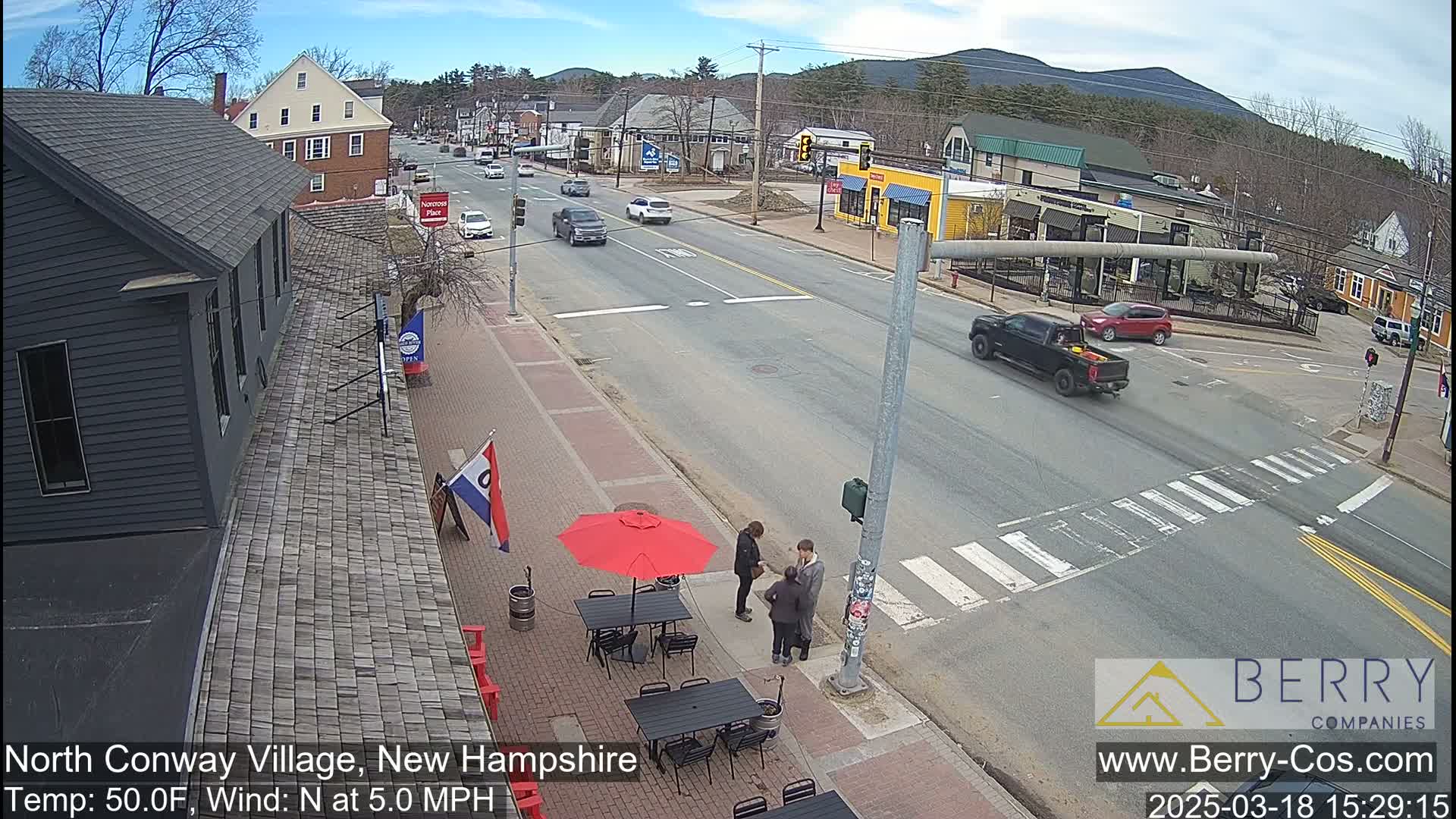 A street scene on a sunny day shows a few cars driving on a road that intersects with a crosswalk where three people are standing near outdoor cafe seating with a red umbrella.