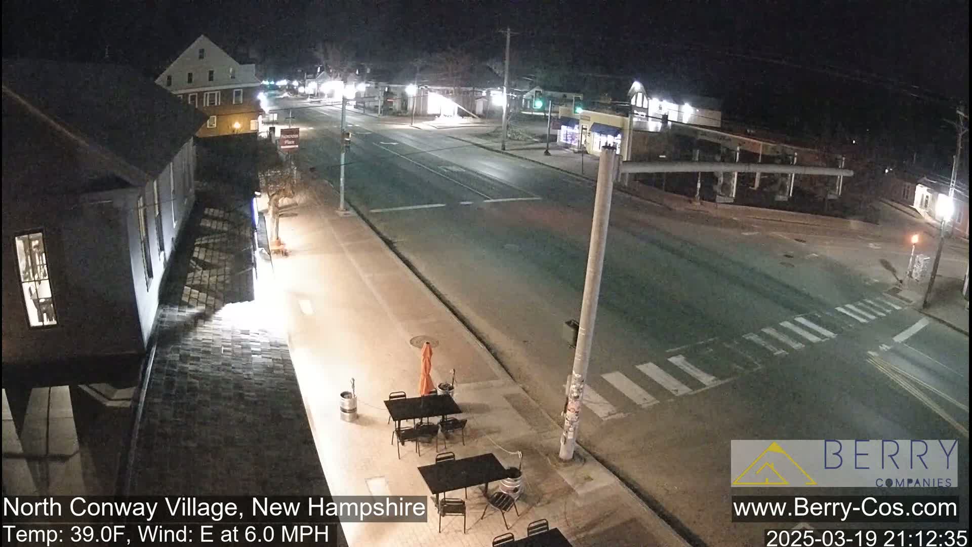 A nighttime street scene shows a mostly empty road with a few buildings and outdoor seating at a restaurant under a clear sky with an east wind.