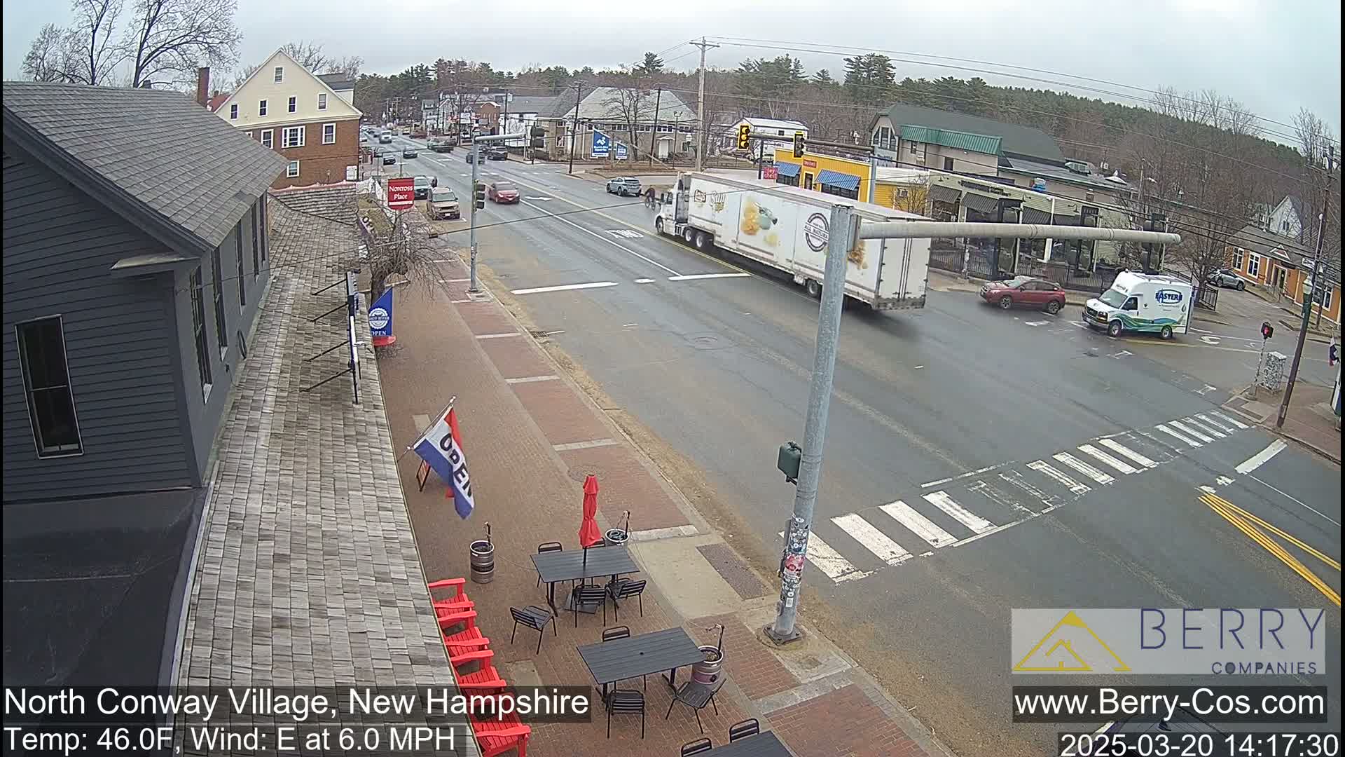 A street scene shows a partly cloudy day with several cars and a large semi-truck at an intersection, with outdoor seating visible at a business along the sidewalk.