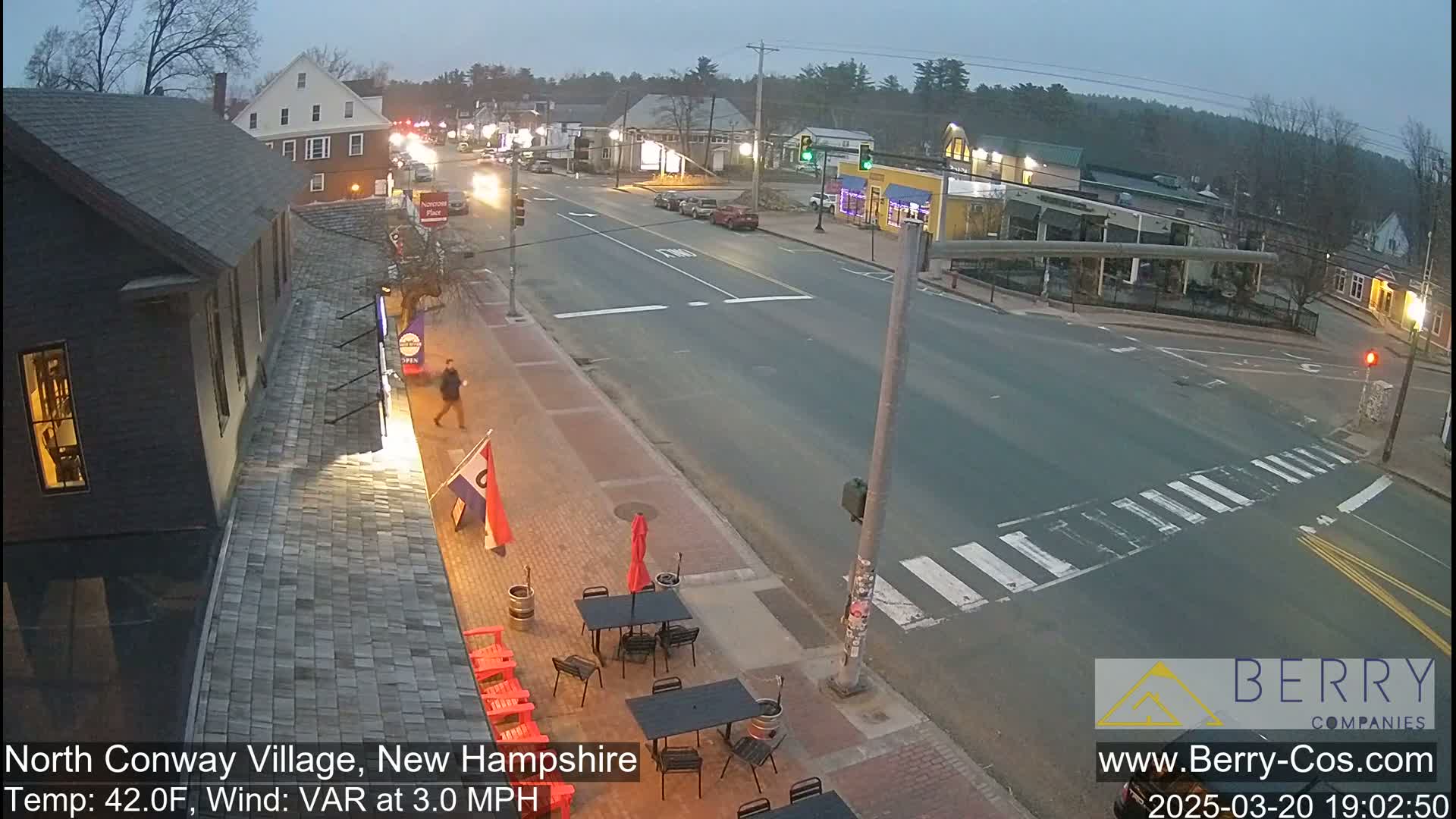 A mostly empty street scene in a village at dusk, showing a few cars and pedestrians near a restaurant with outdoor seating under a mostly clear sky, with a light wind.