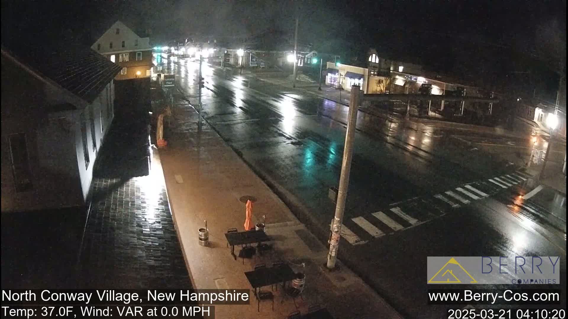 A rain-slicked street scene at night shows a mostly empty roadway with a few buildings visible, and some outdoor patio furniture.