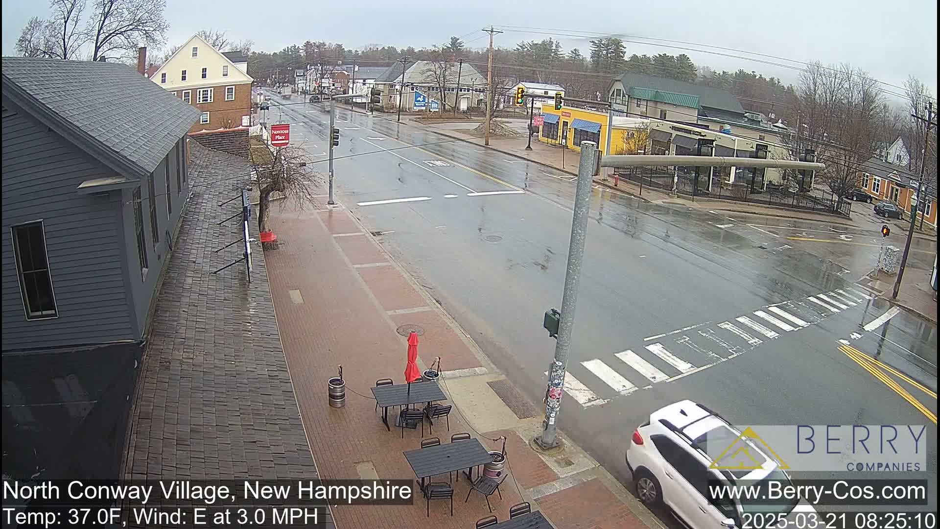 A wet street scene shows a mostly empty intersection in a small town with some buildings, a few cars, and outdoor cafe seating.