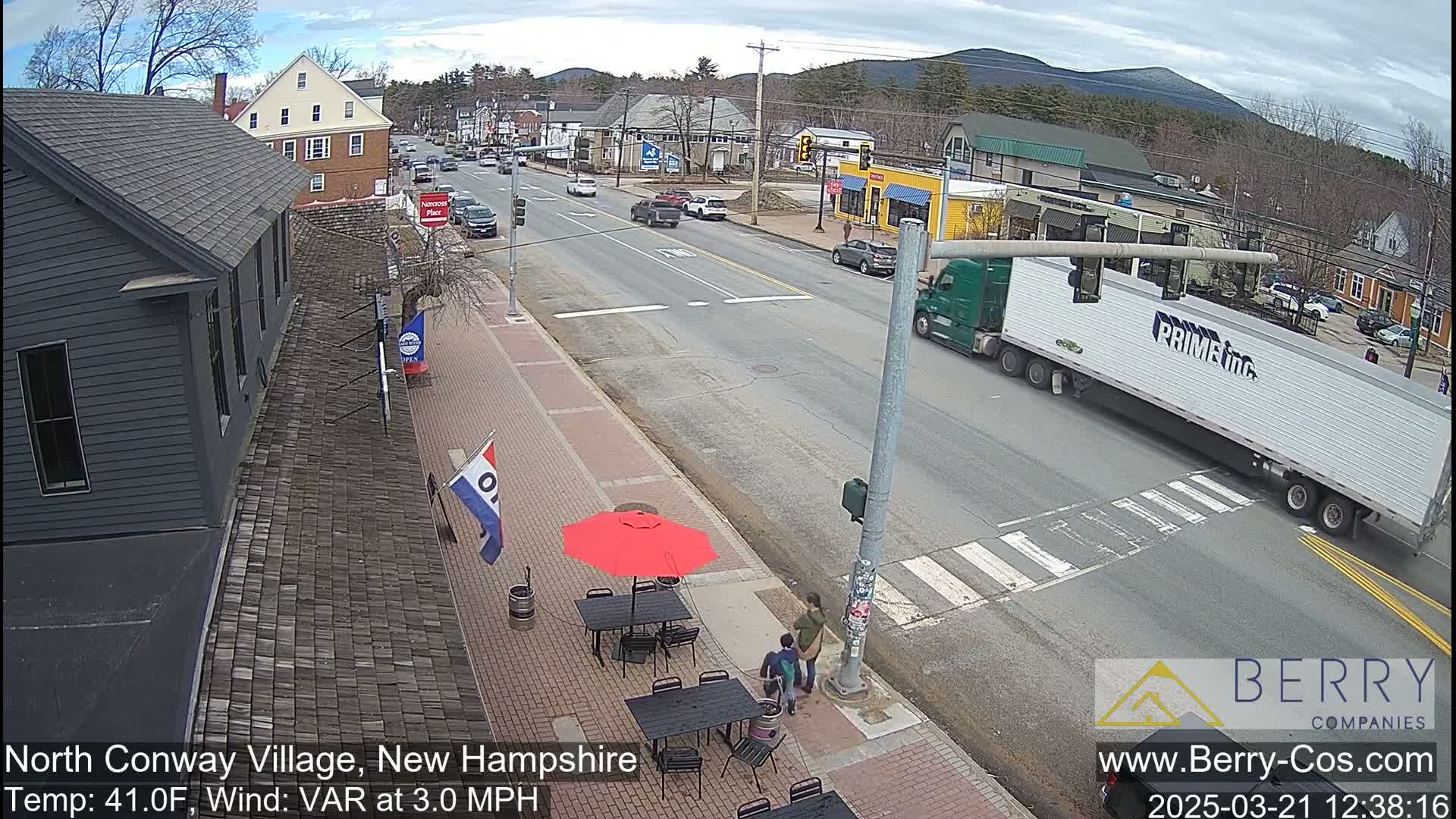 A street scene on a partly sunny day shows a main road with cars and a large semi-truck at an intersection, with a smaller side street and buildings, including a dark-colored building with a red umbrella outside, and mountains in the distance.