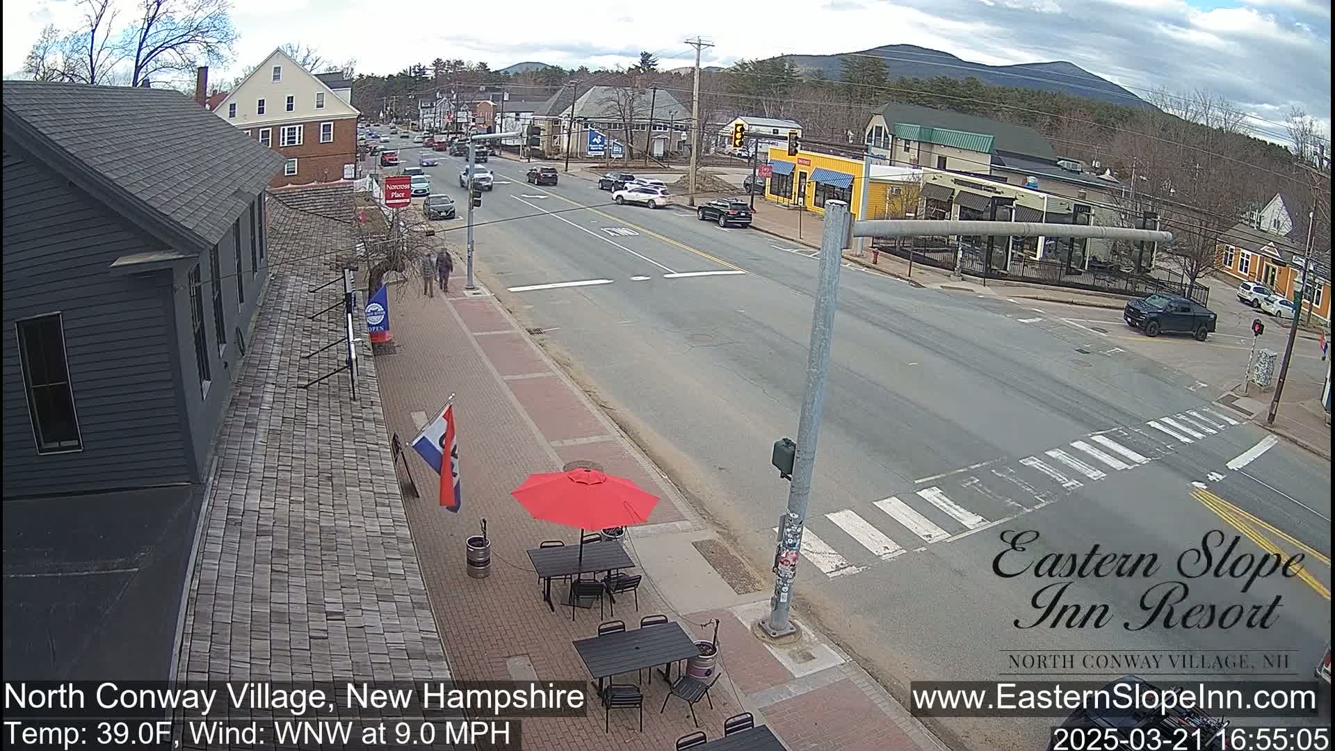 A street scene in a village shows a four-way intersection with several cars and pedestrians, under a partly cloudy sky.