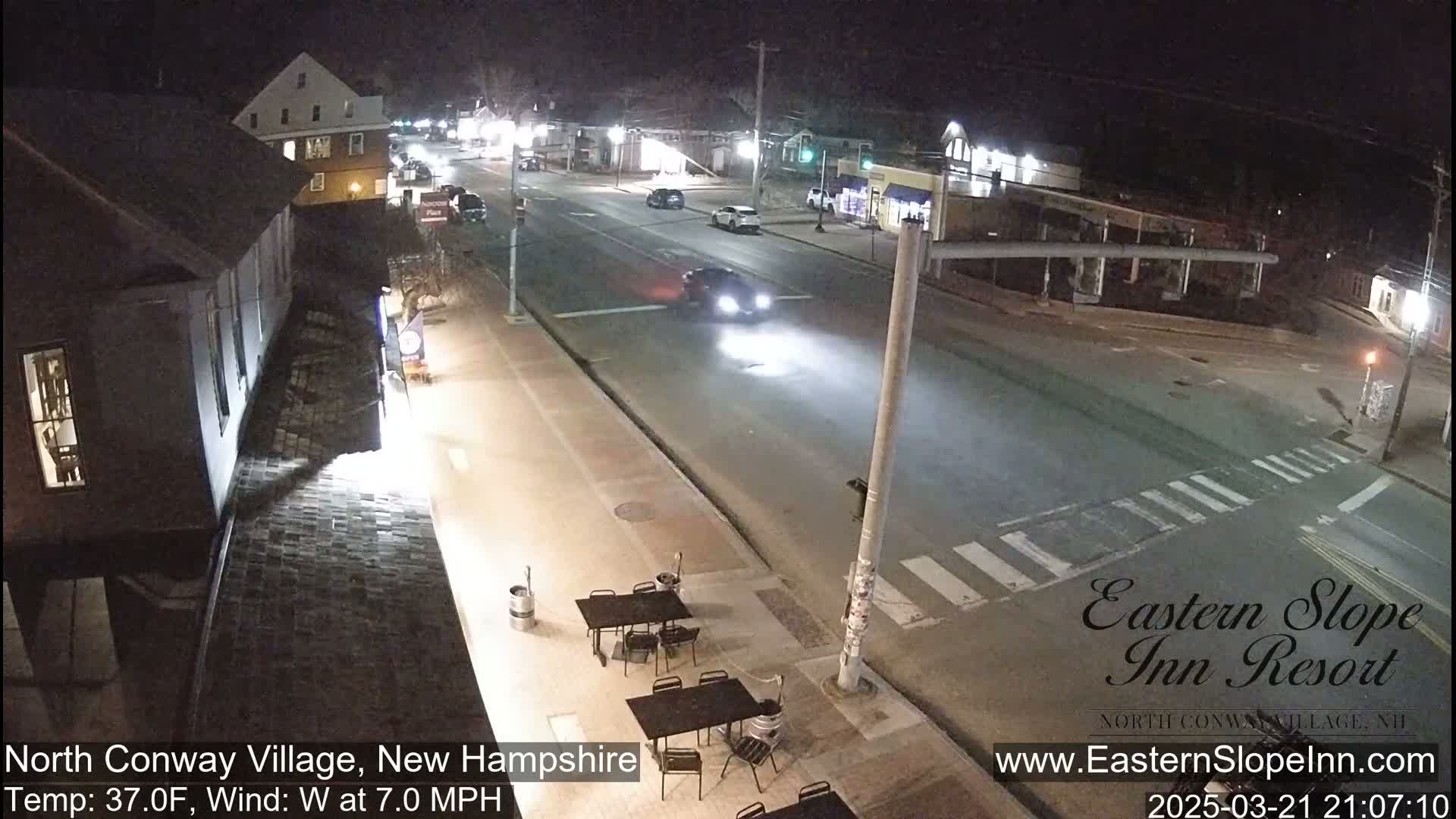 A nighttime street scene shows a few cars driving on a road that intersects with another road, with outdoor seating visible in front of a building under a clear sky, and a 37°F temperature with a 7 mph west wind.