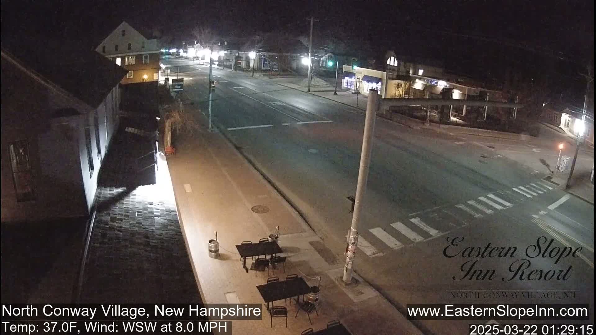 A nighttime street scene shows a mostly empty road intersecting with a crosswalk,  a few outdoor tables and chairs, and buildings under partly cloudy conditions;  the temperature is 37°F with a west-southwest wind at 8 mph.