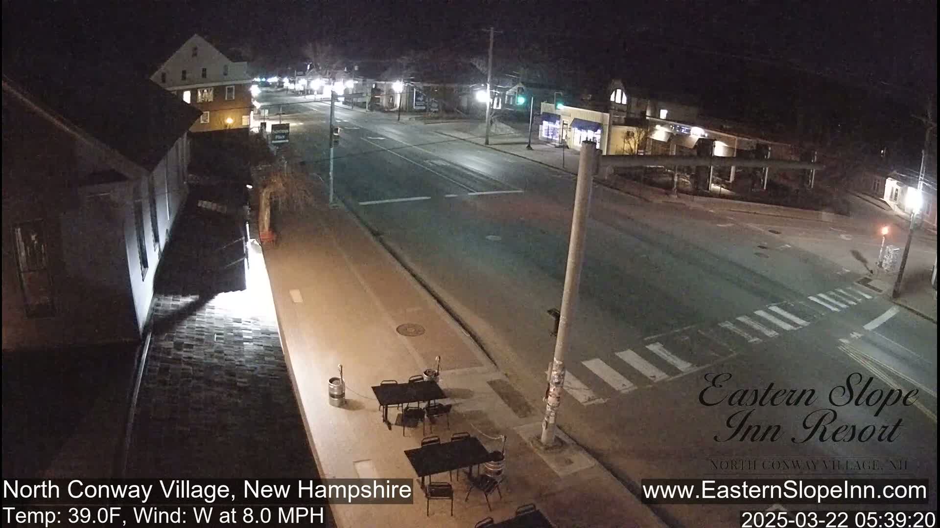 A nighttime, mostly empty street scene with a few buildings and outdoor seating visible, under clear skies and a 39°F temperature with an 8 mph west wind.