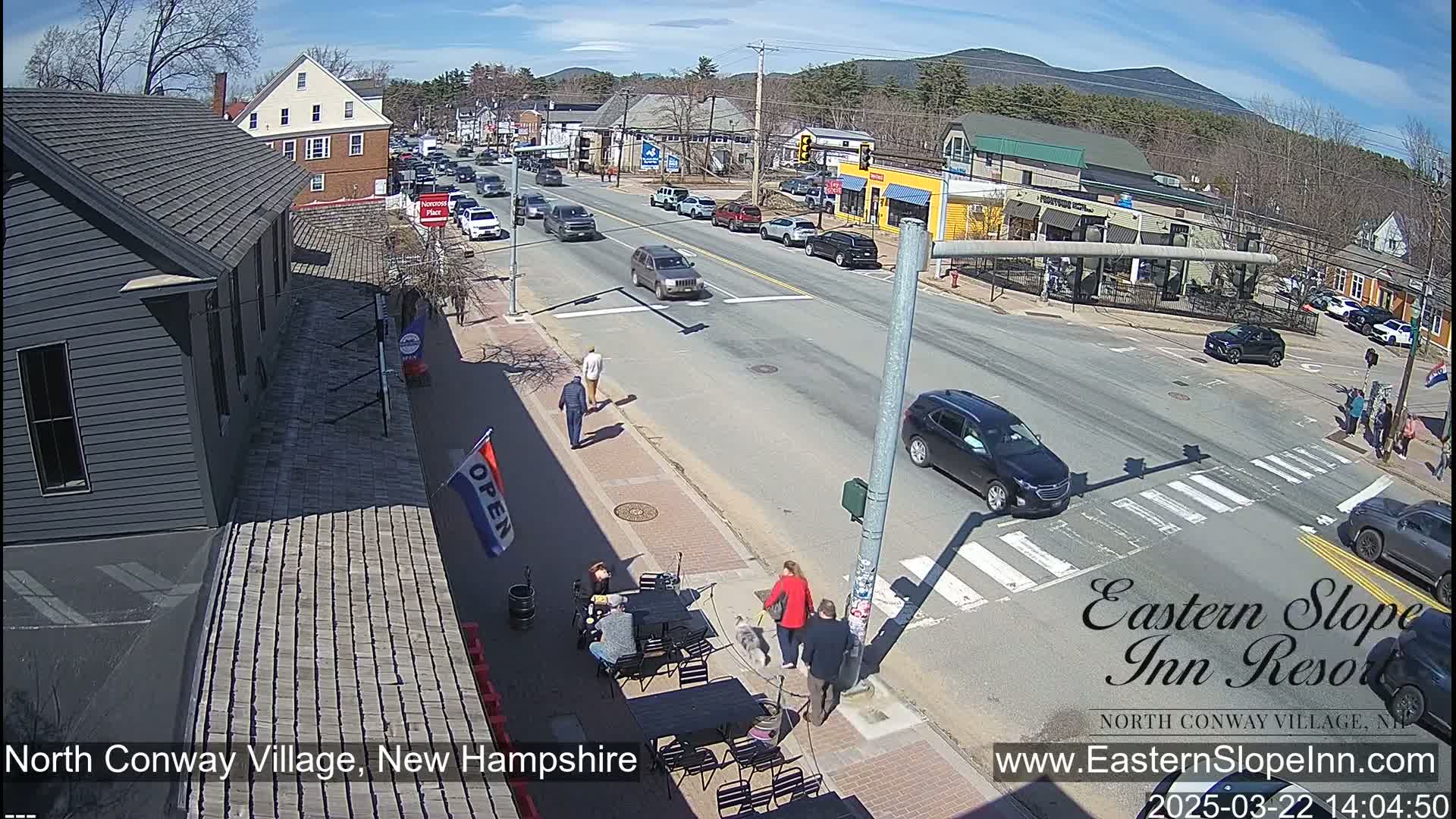 A sunny street scene in a small town shows cars at a crossroads, pedestrians walking on sidewalks, and outdoor seating at a cafe.