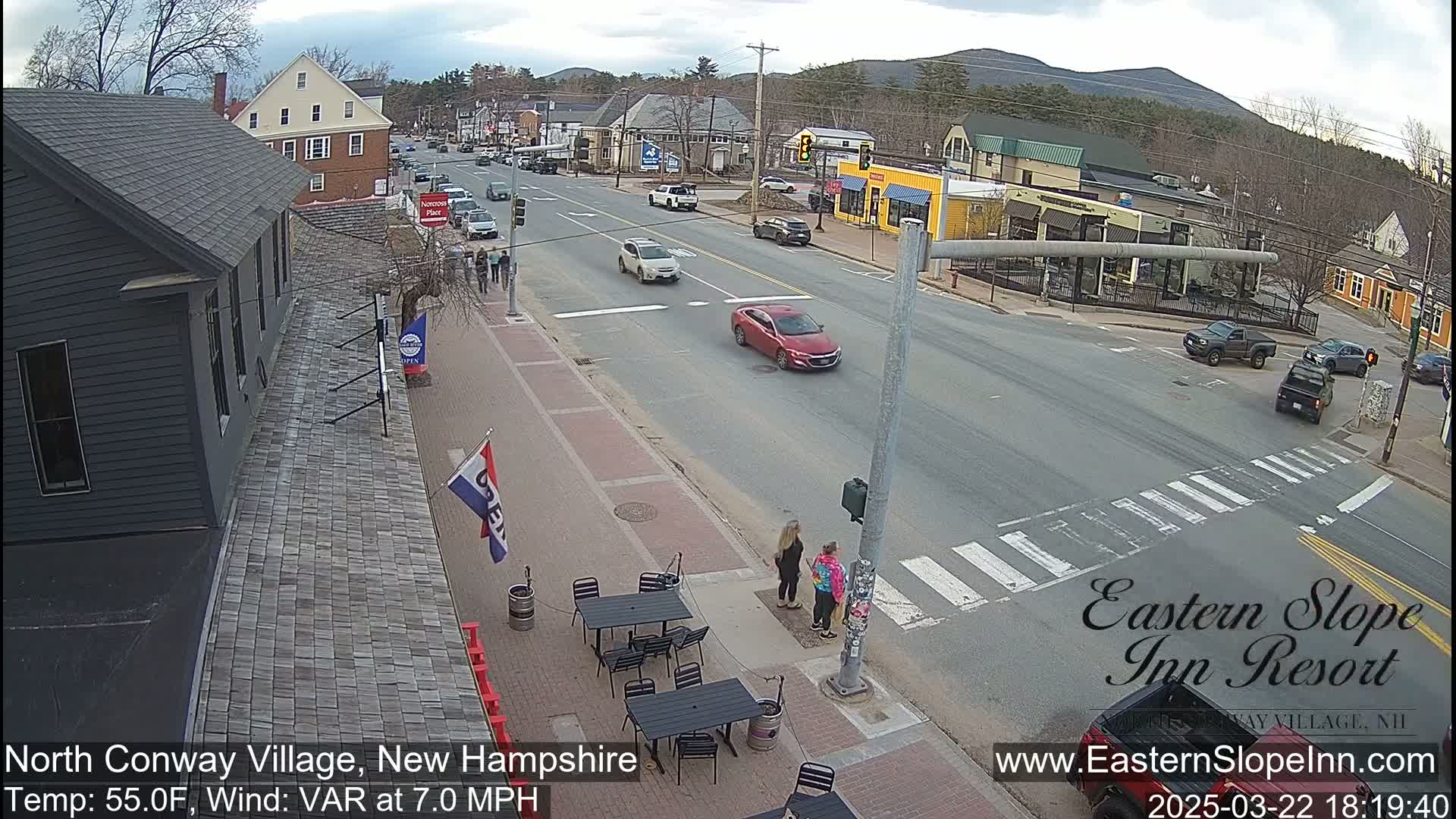 A mostly sunny, street-level view of a town intersection with several cars, pedestrians, and outdoor seating at a restaurant, under a partly cloudy sky.