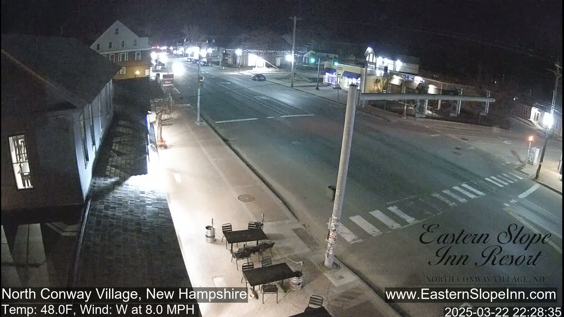 A nighttime street scene shows a mostly empty road with a few cars, buildings, and outdoor patio furniture.