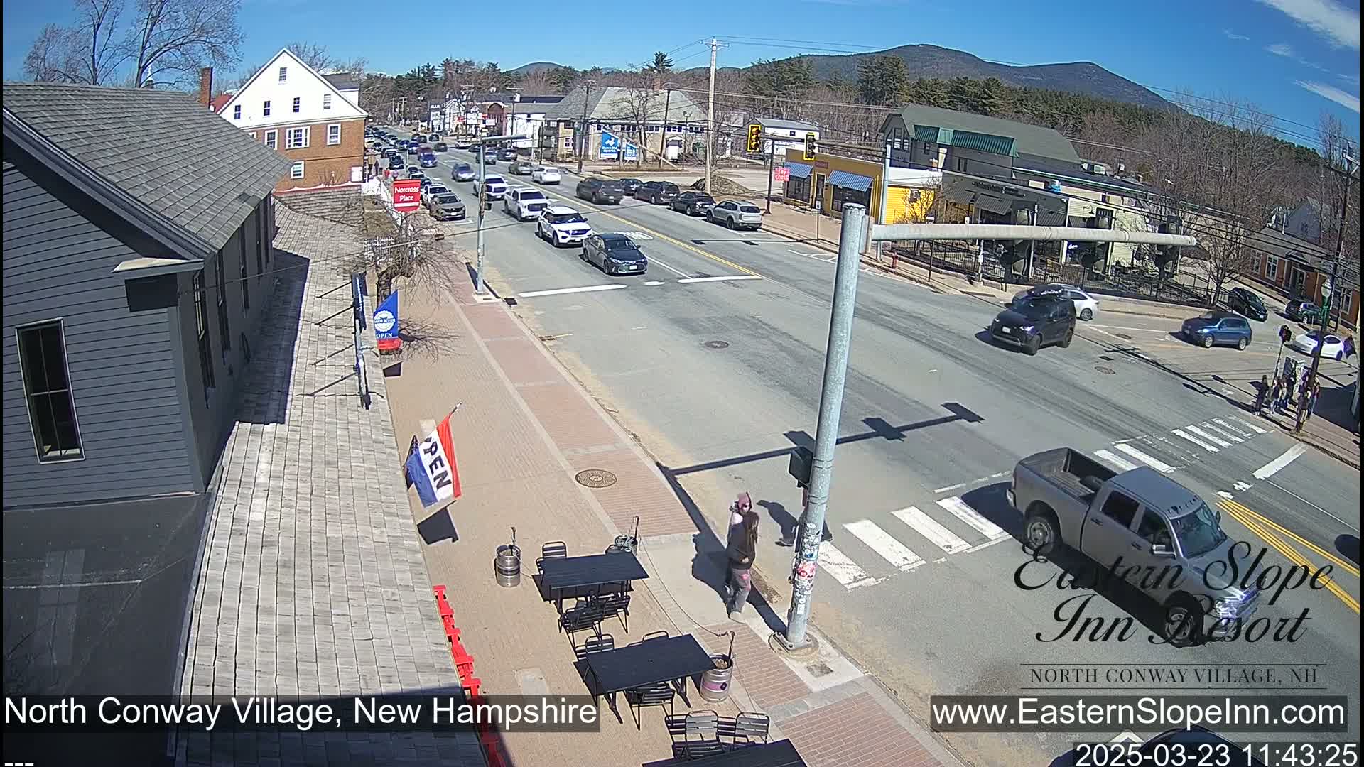 A sunny street scene shows several cars driving on a road in front of buildings, with outdoor seating visible at a business on the left.