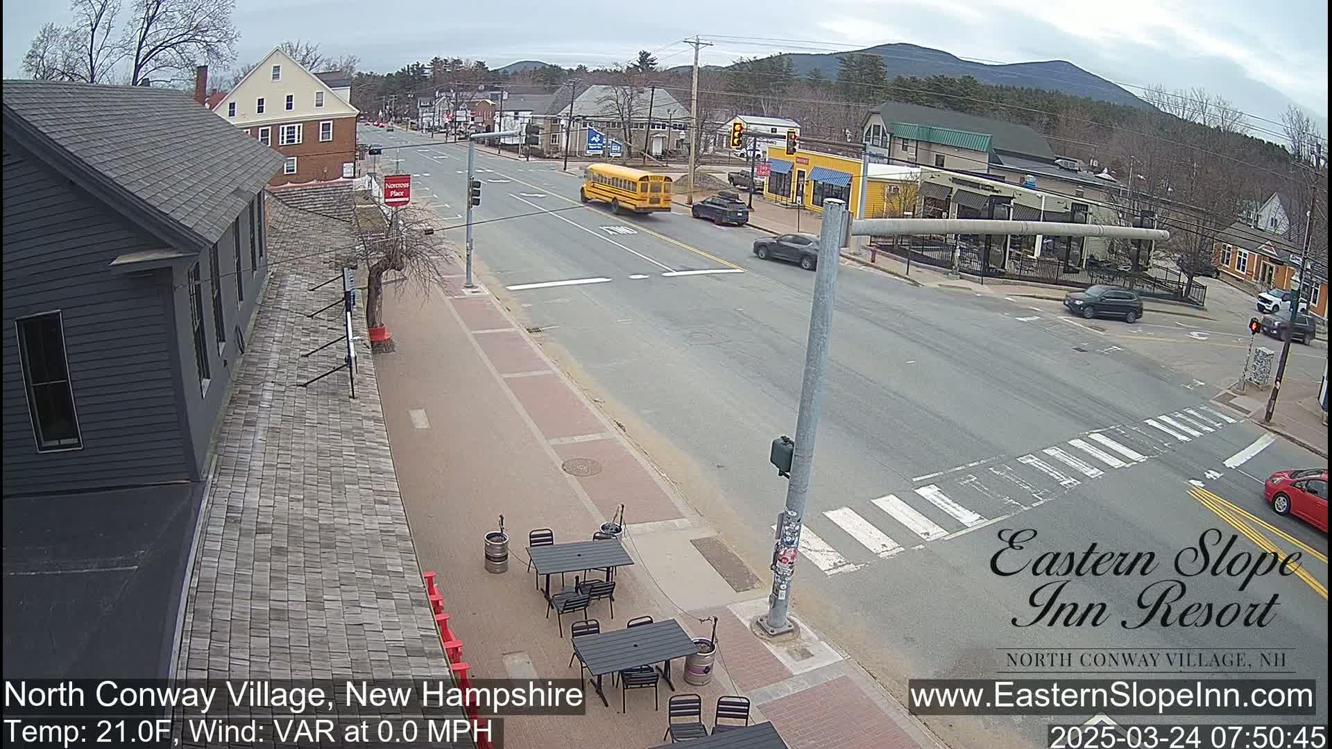 A mostly empty street scene in a village, under a cloudy sky, features a school bus, several cars, and outdoor seating at a restaurant.